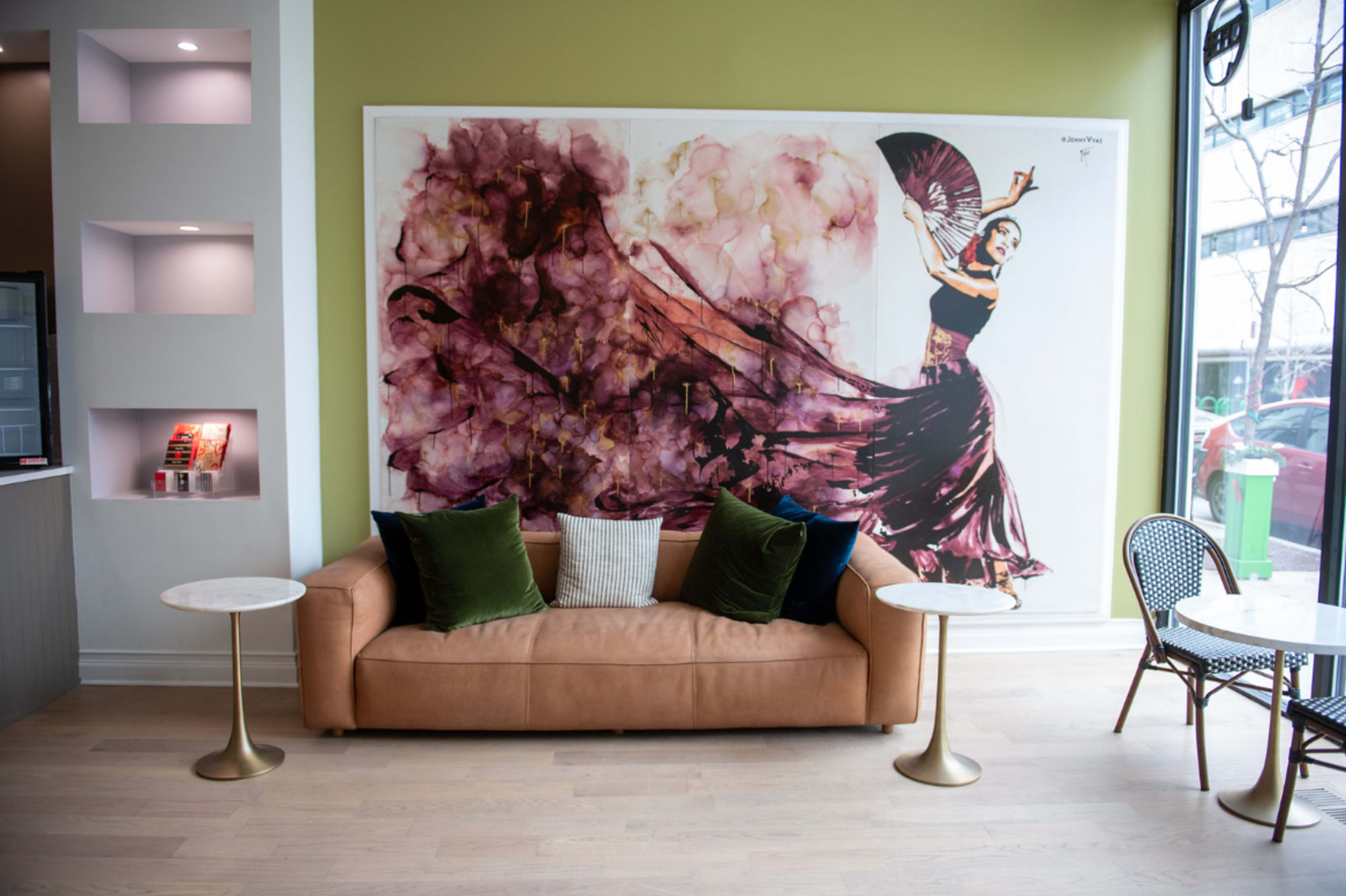 A modern seating area featuring a brown sofa with green and white cushions, flanked by small round tables and a large wall mural depicting a dancer.