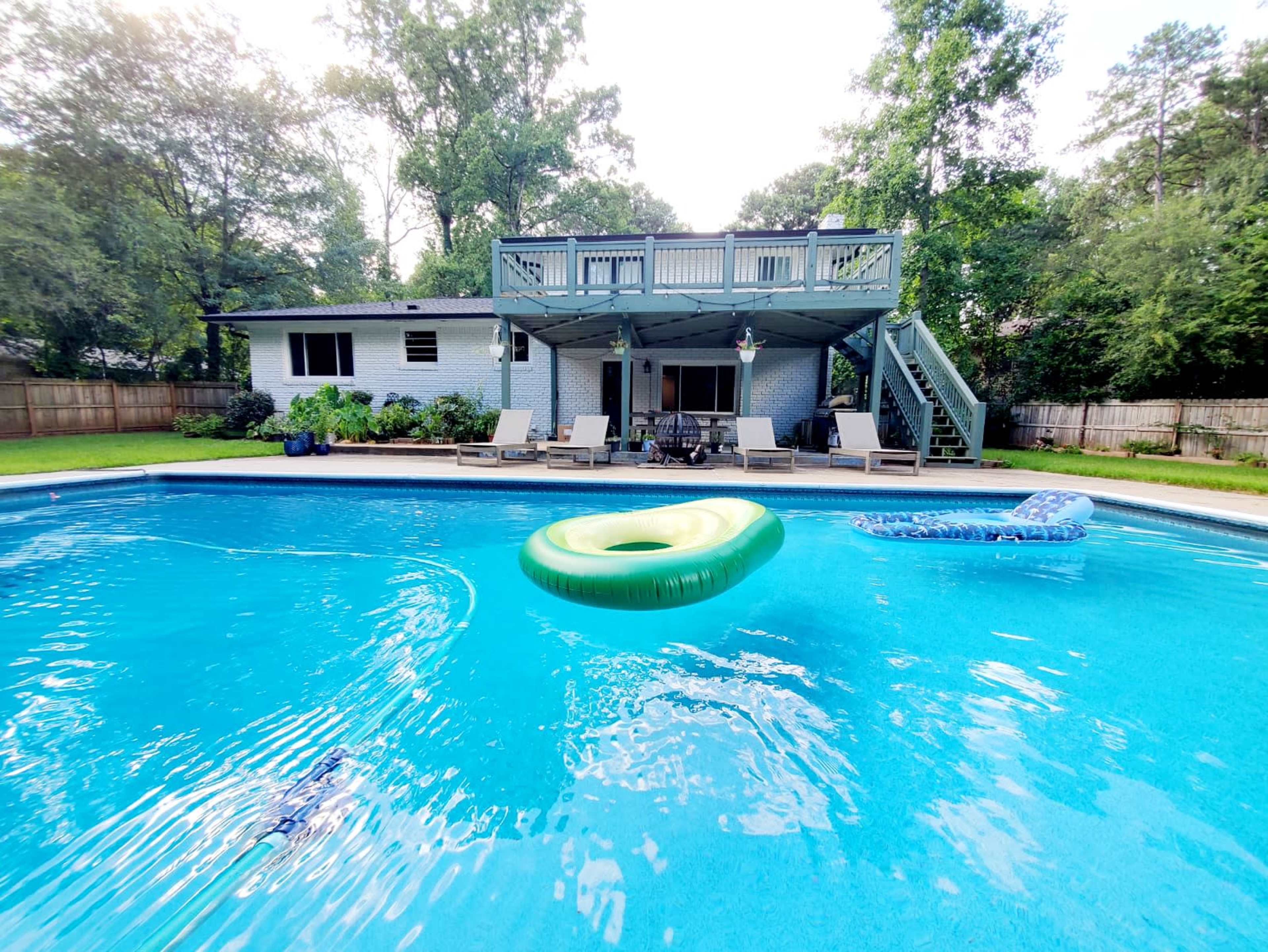 A bright blue swimming pool features a green inflatable float, with a two-story deck and a house visible in the background surrounded by trees.