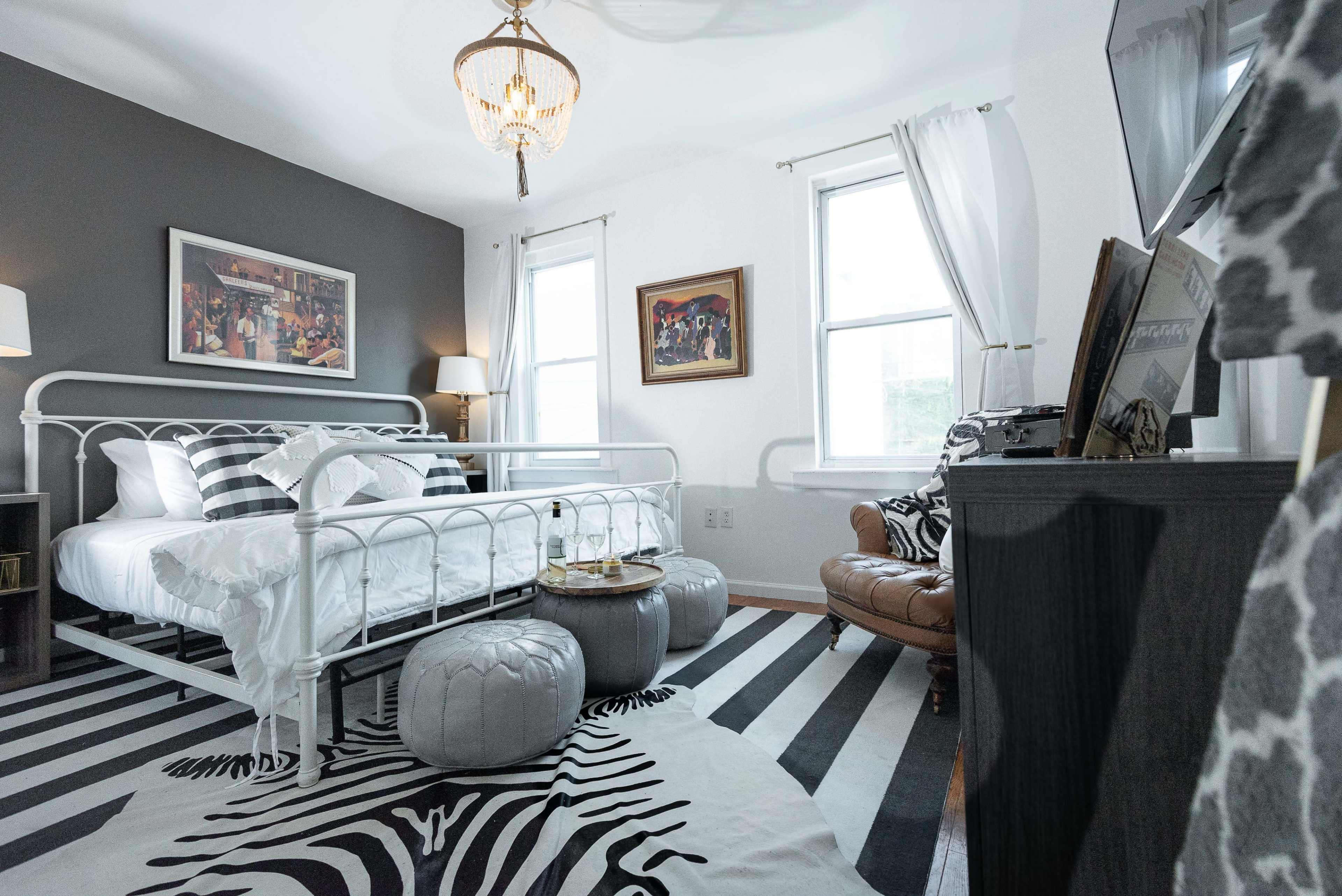 The bedroom features a white metal bed with black and white bedding, a leather armchair, and a striped rug.