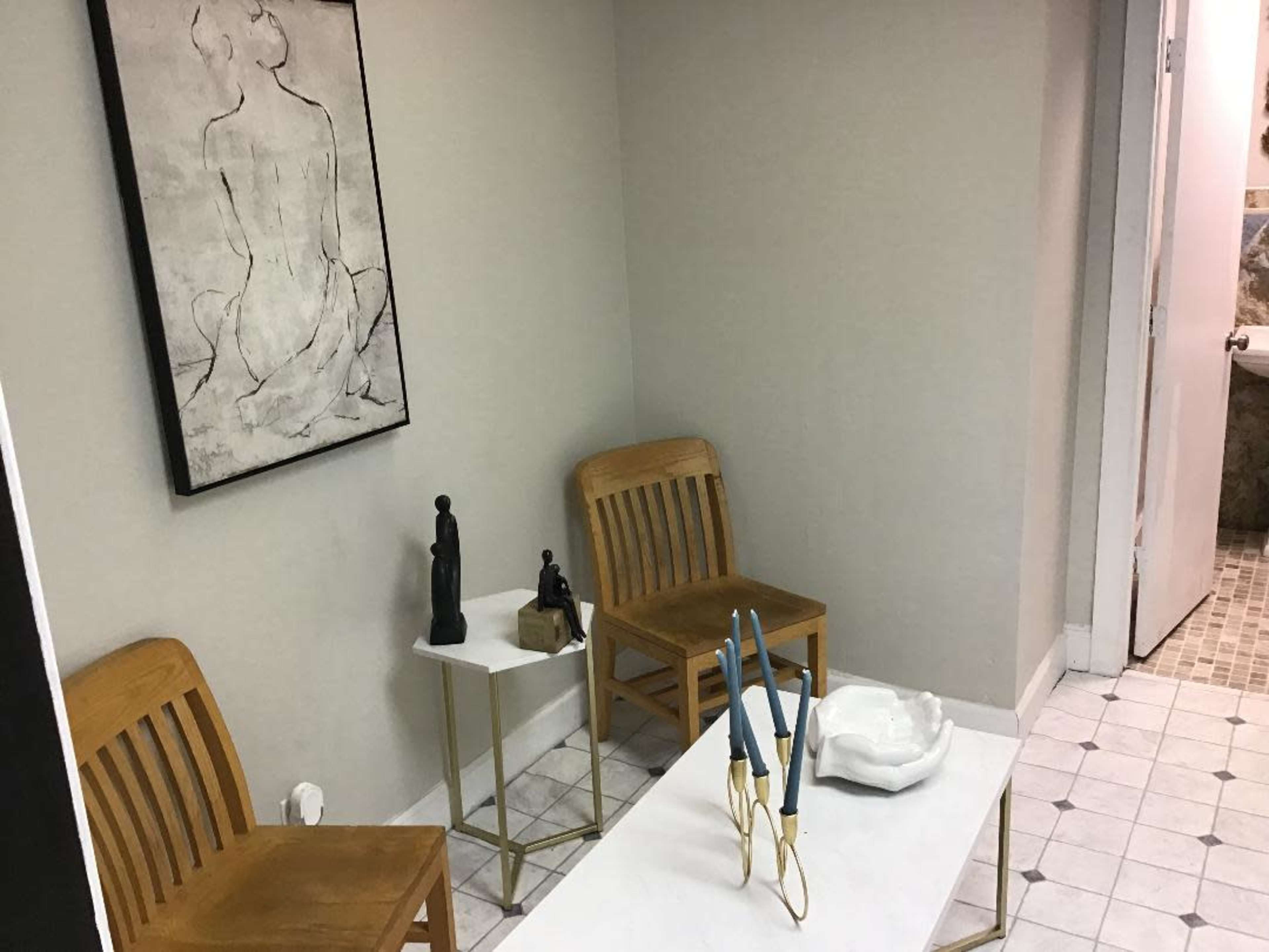 The image shows a small room with two wooden chairs, a side table, and various decorative objects, including candles and a sculpture, against a light-colored wall.