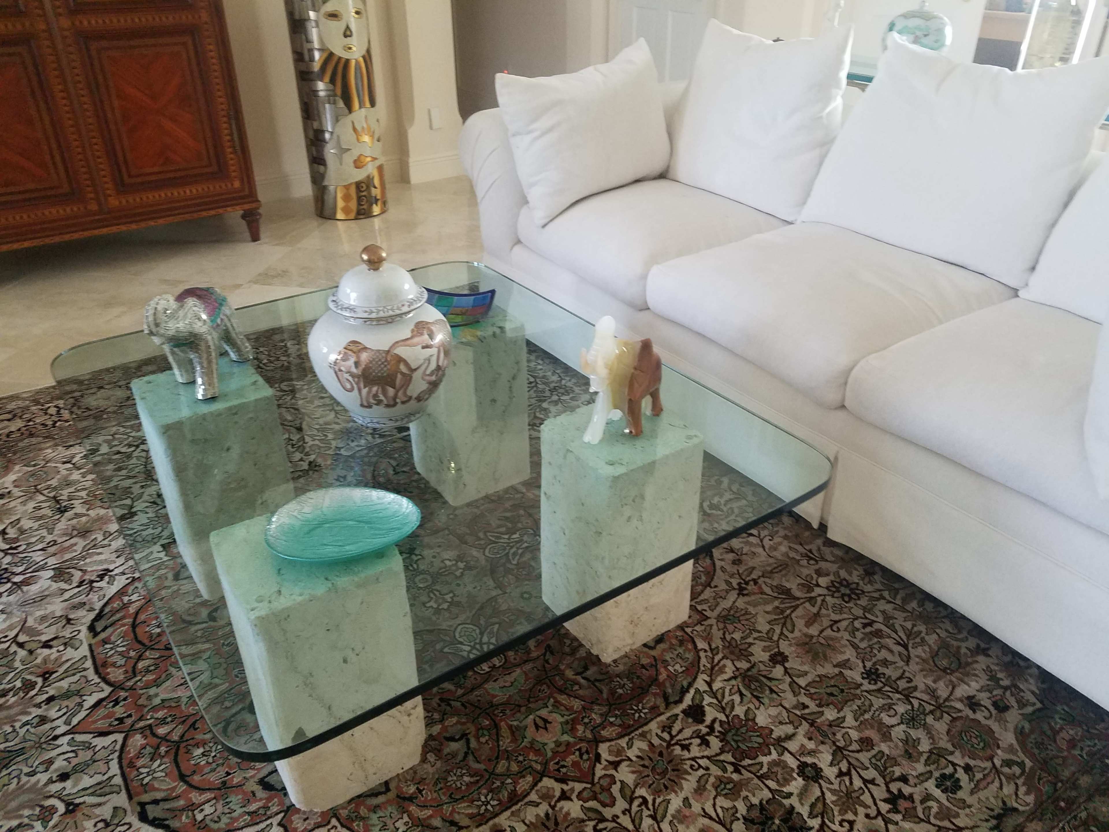 A glass coffee table with stone legs is placed in front of a white sofa, surrounded by various decorative items, including a porcelain jar and two small elephant figures, on a patterned rug.