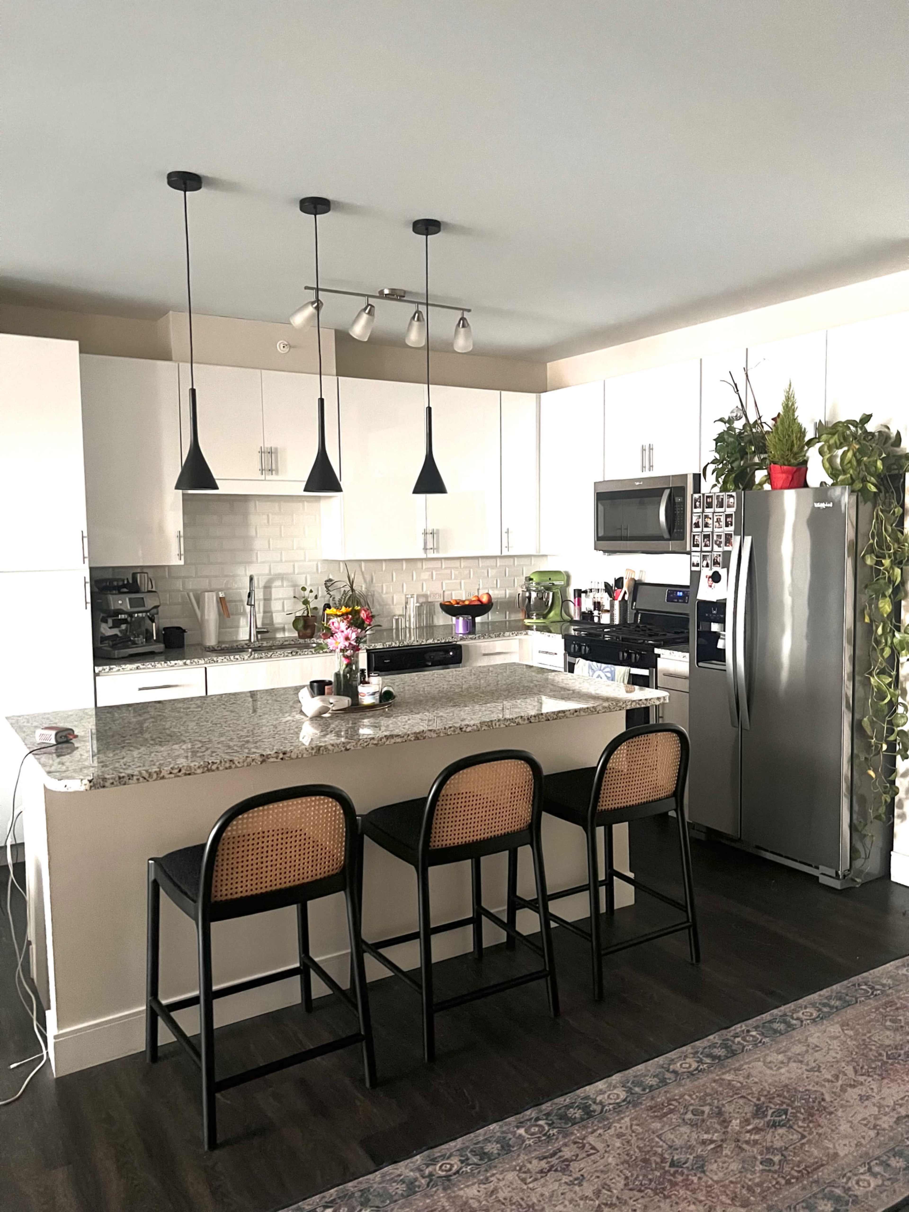 A modern kitchen features a central island with four black stools, white cabinets, granite countertops, and stainless steel appliances.