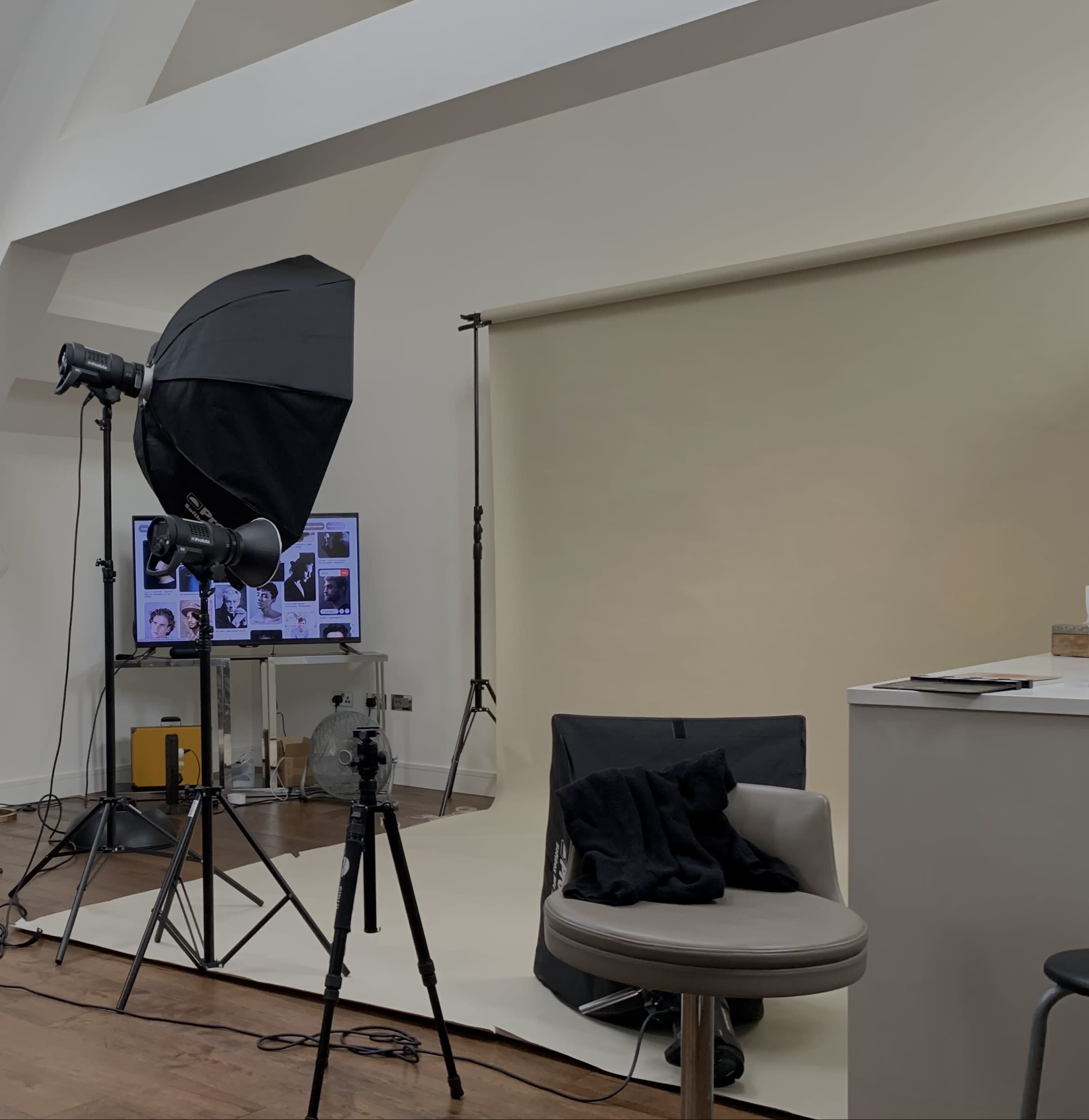 A photography studio setup featuring a backdrop, lighting equipment, and a chair, with a computer screen displaying images in the background.