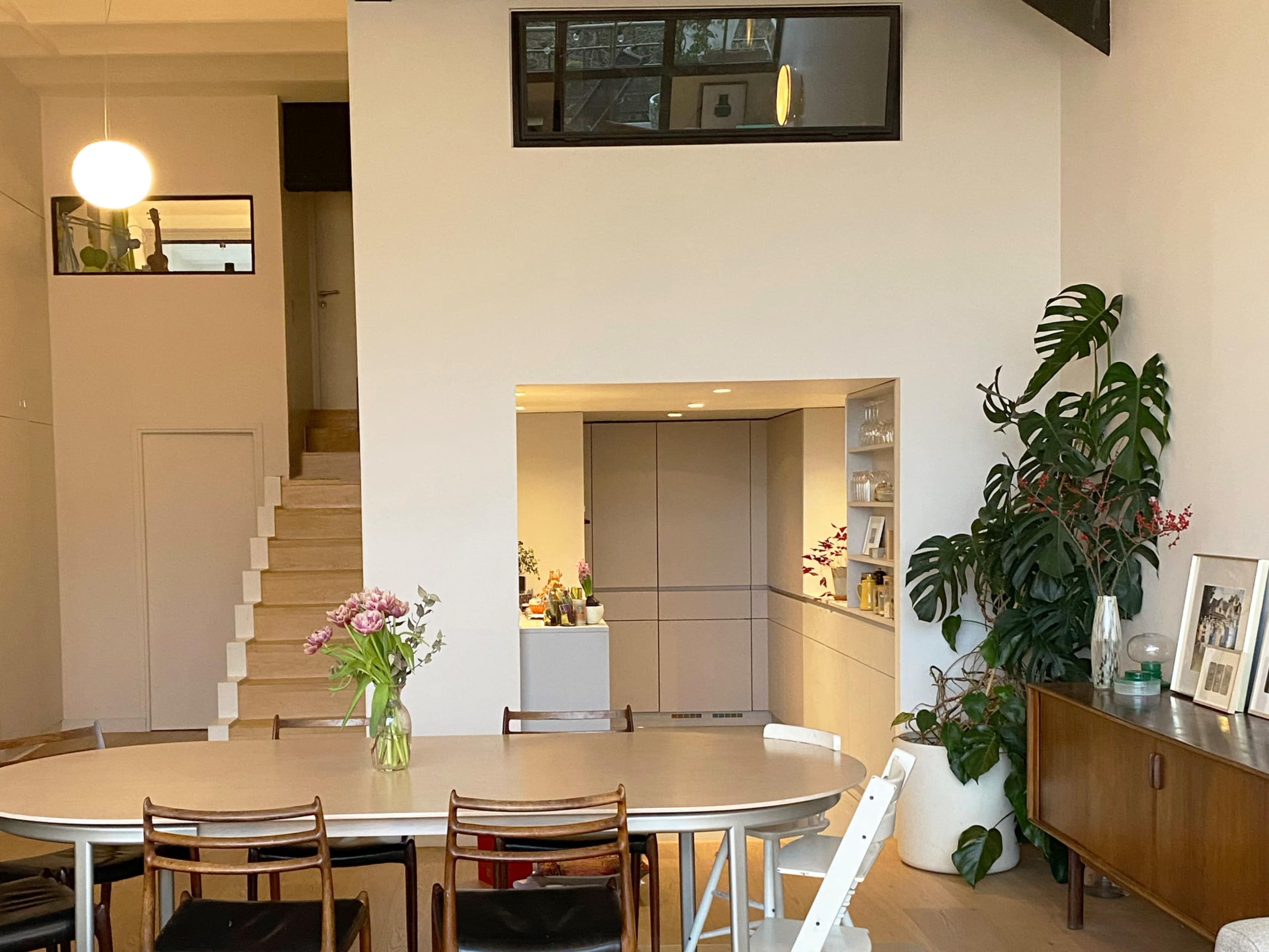 The image shows a modern interior with a dining table surrounded by chairs, a staircase leading to an upper level, and a kitchen area visible in the background, adorned with plants and decorative items.