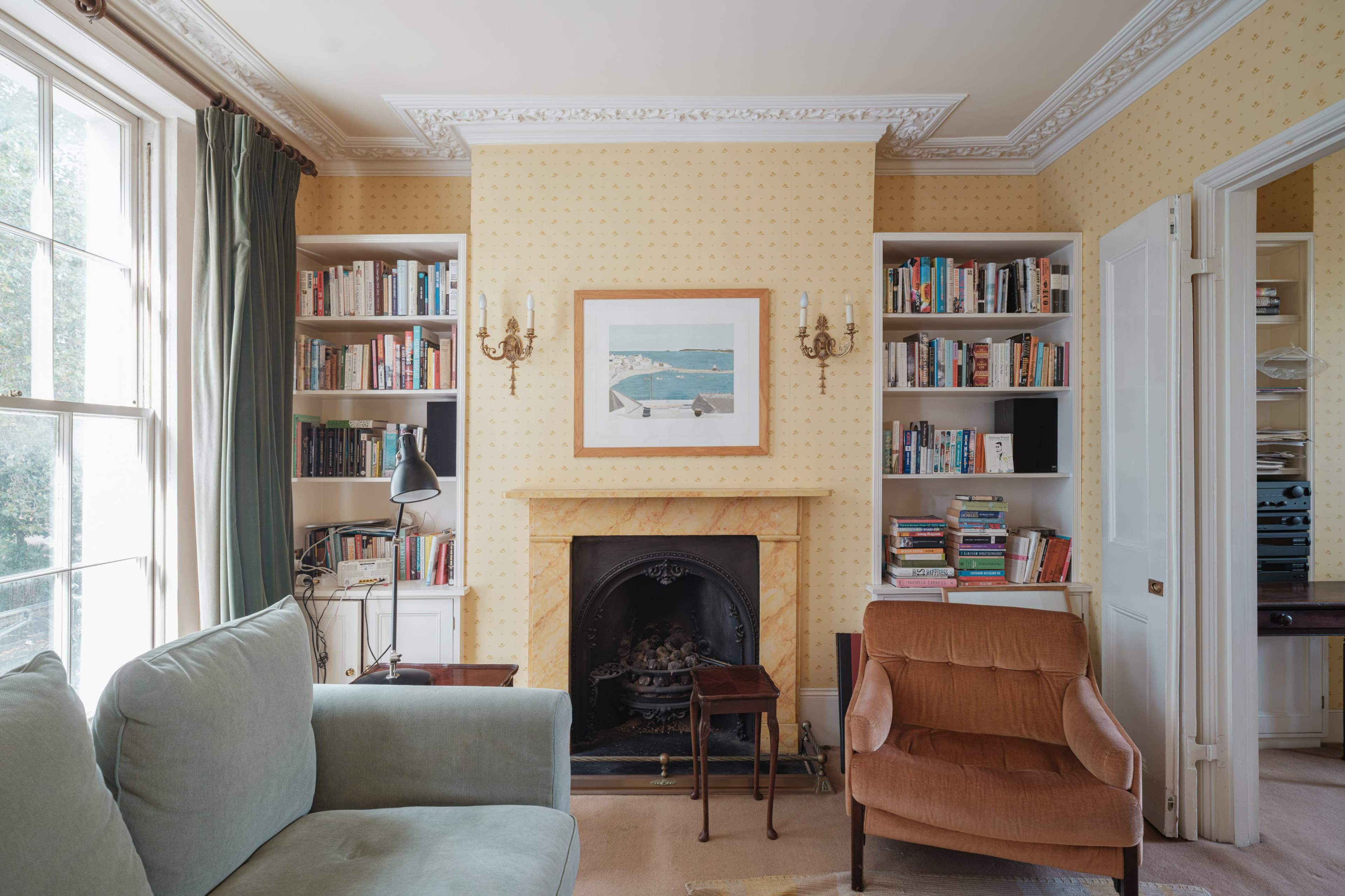 A cozy living room features a fireplace surrounded by bookshelves, a framed painting on the wall, and a sofa paired with an armchair.