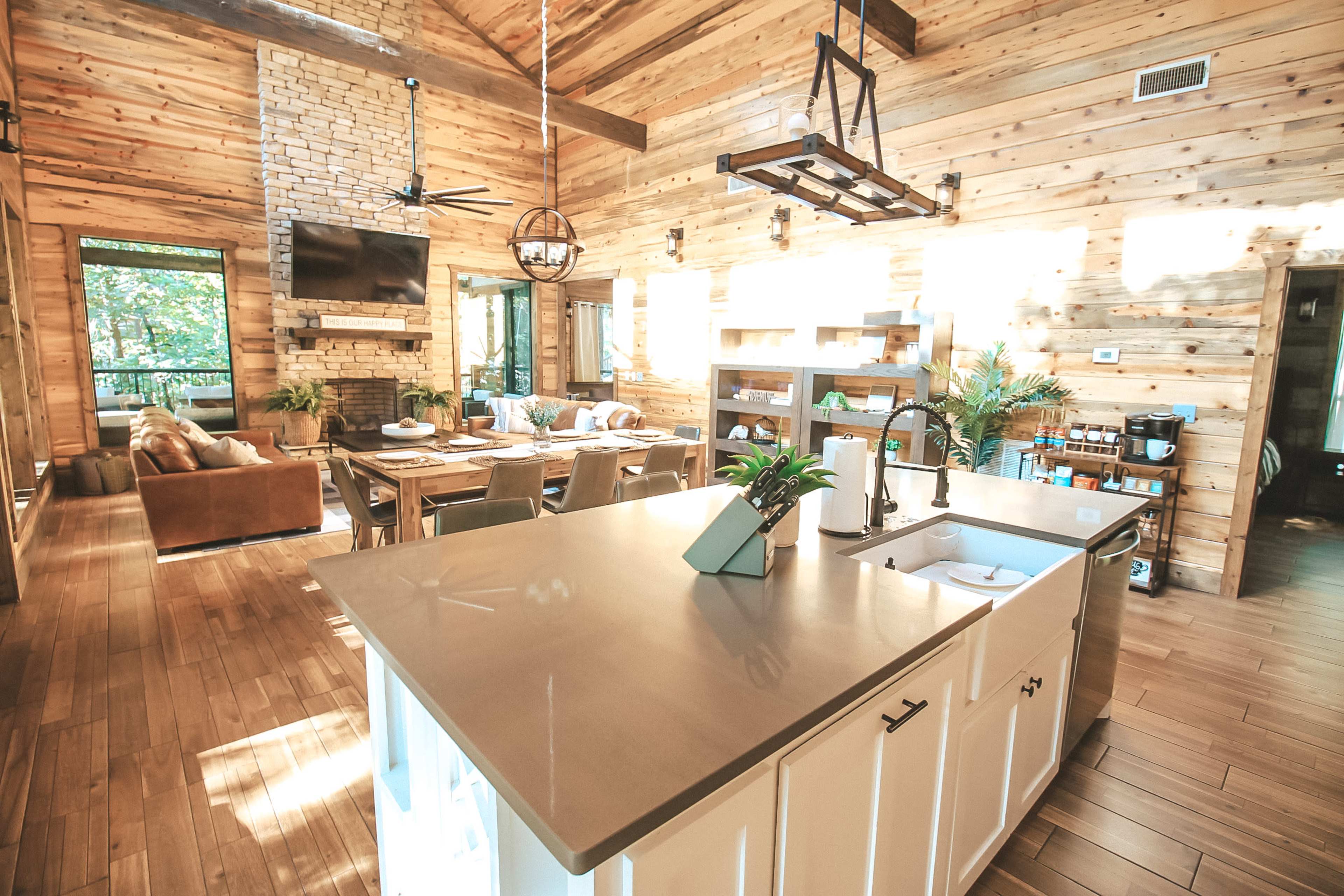 A spacious wood-paneled cabin interior with an open kitchen, dining area, and living space featuring a stone fireplace and large windows.
