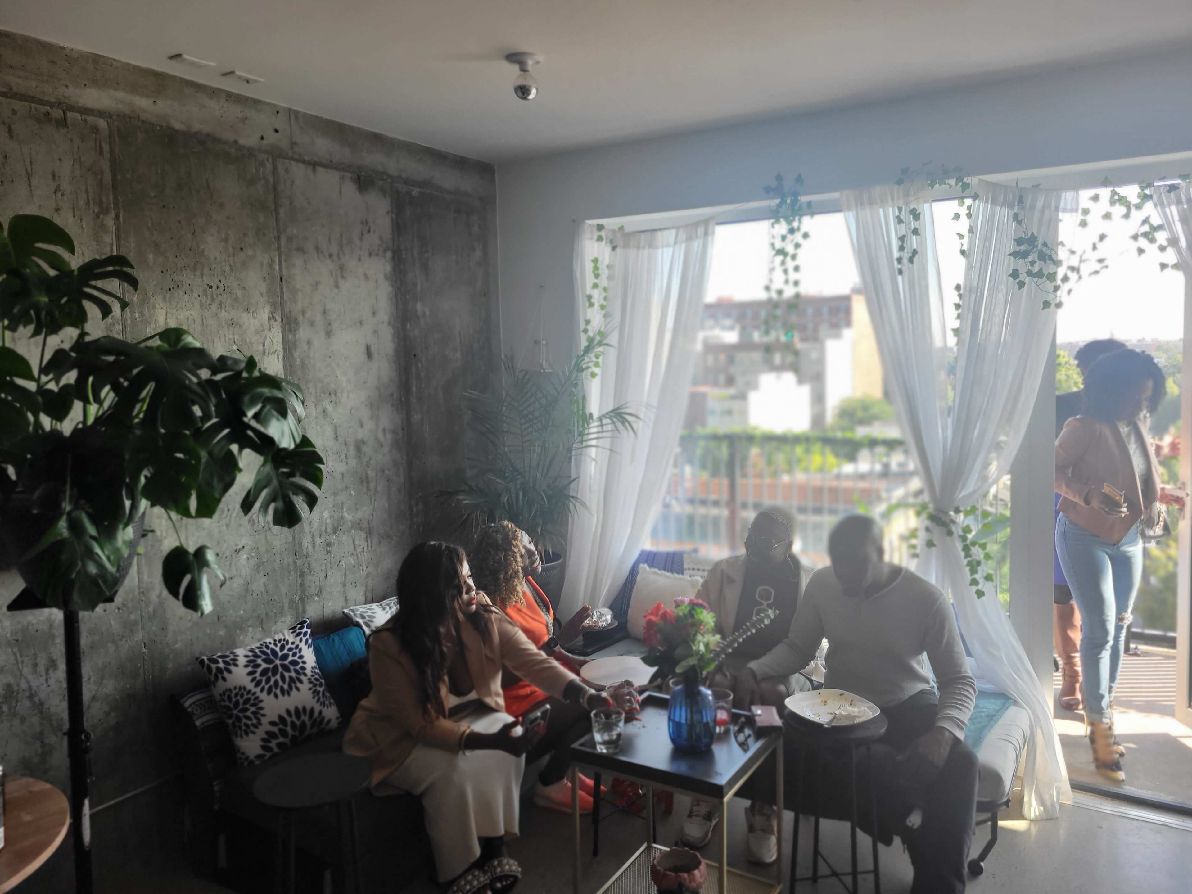 A group of people sit and stand around a table in a stylish room with large windows overlooking a cityscape.