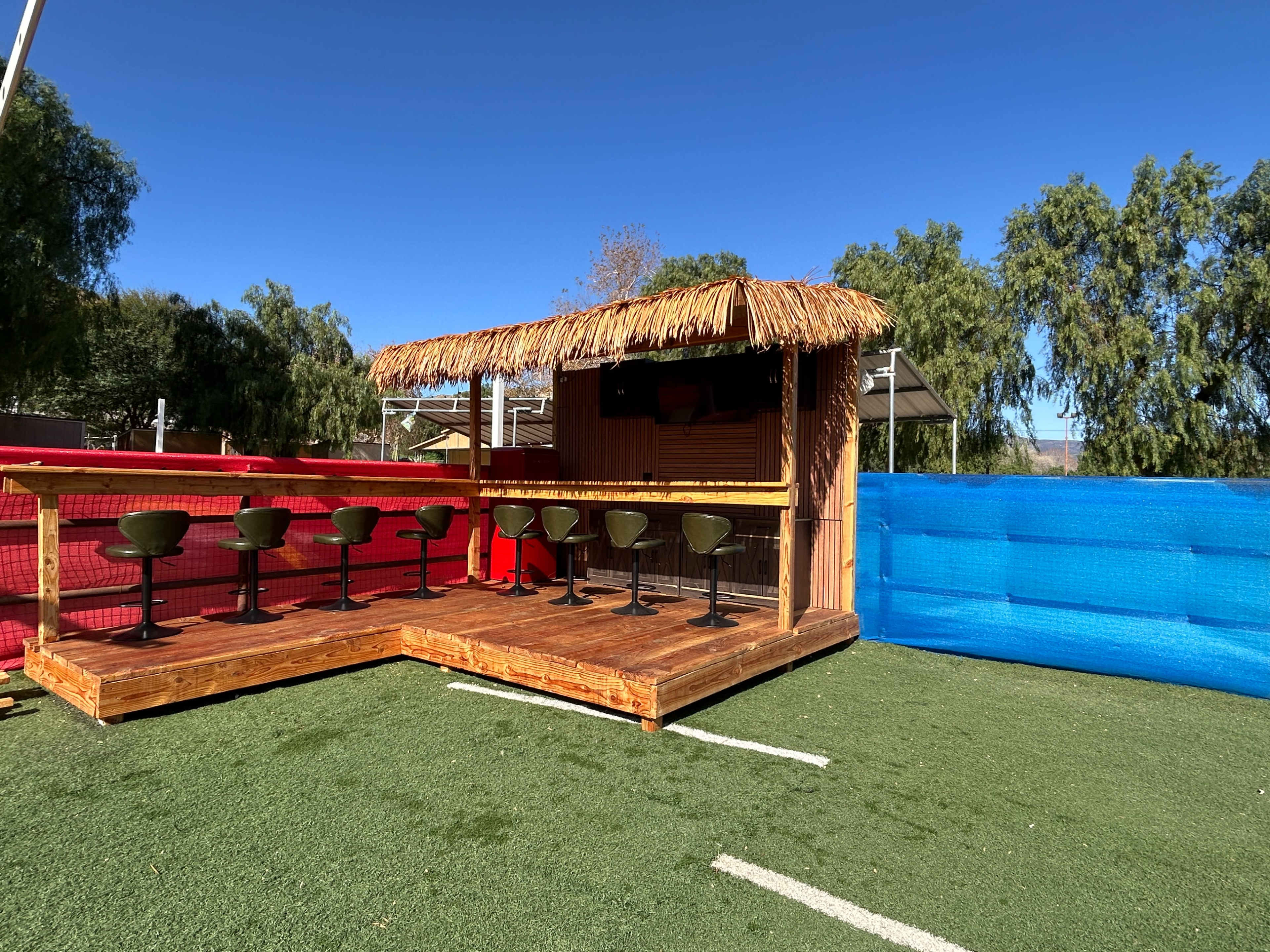 The image shows a wooden outdoor bar area with a thatched roof, lined with bar stools, set against a backdrop of green grass and colorful fencing.