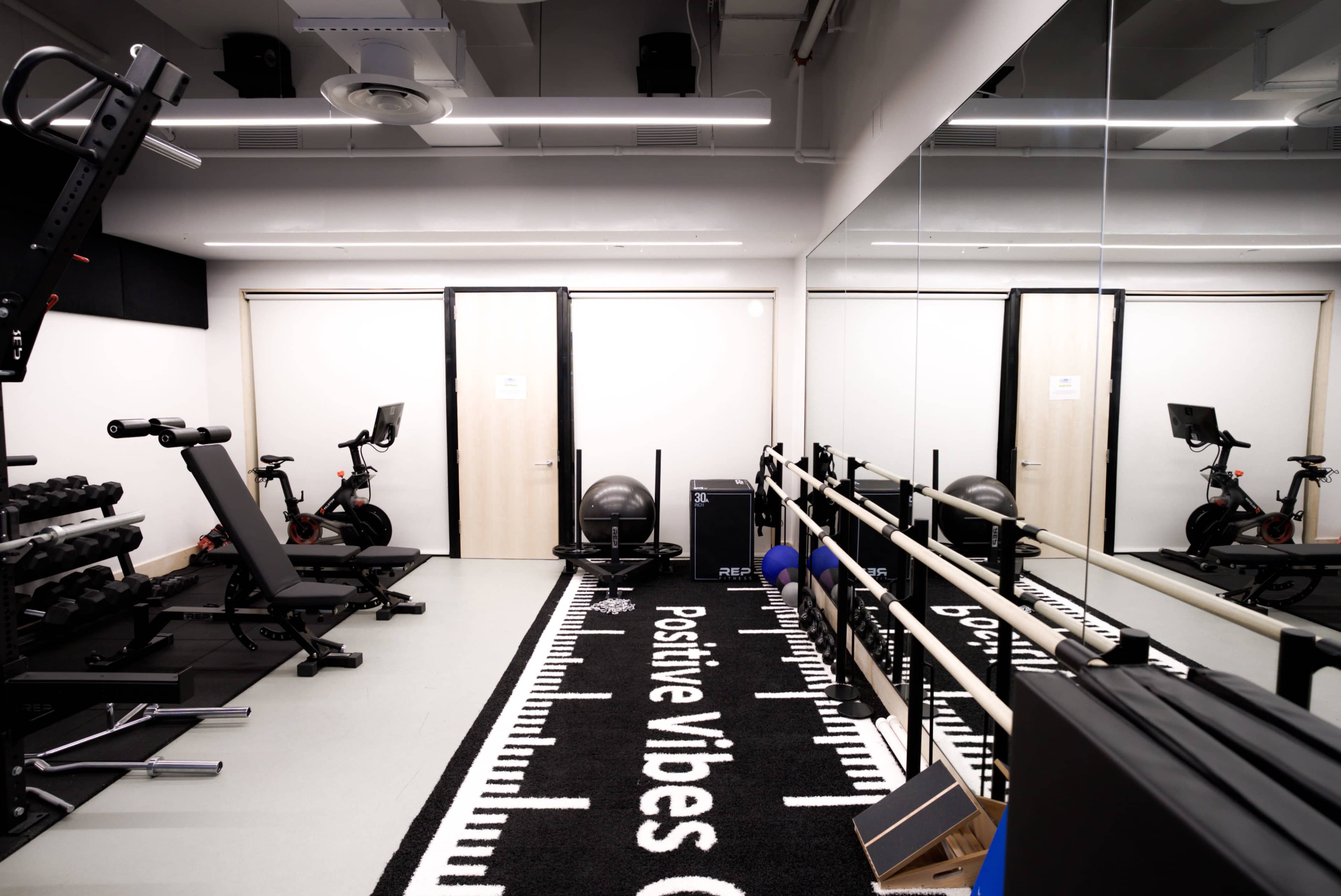 A modern gym space equipped with weights, exercise machines, a stability ball, and mirrors, featuring a carpet with the phrase "Positive Vibes Only."