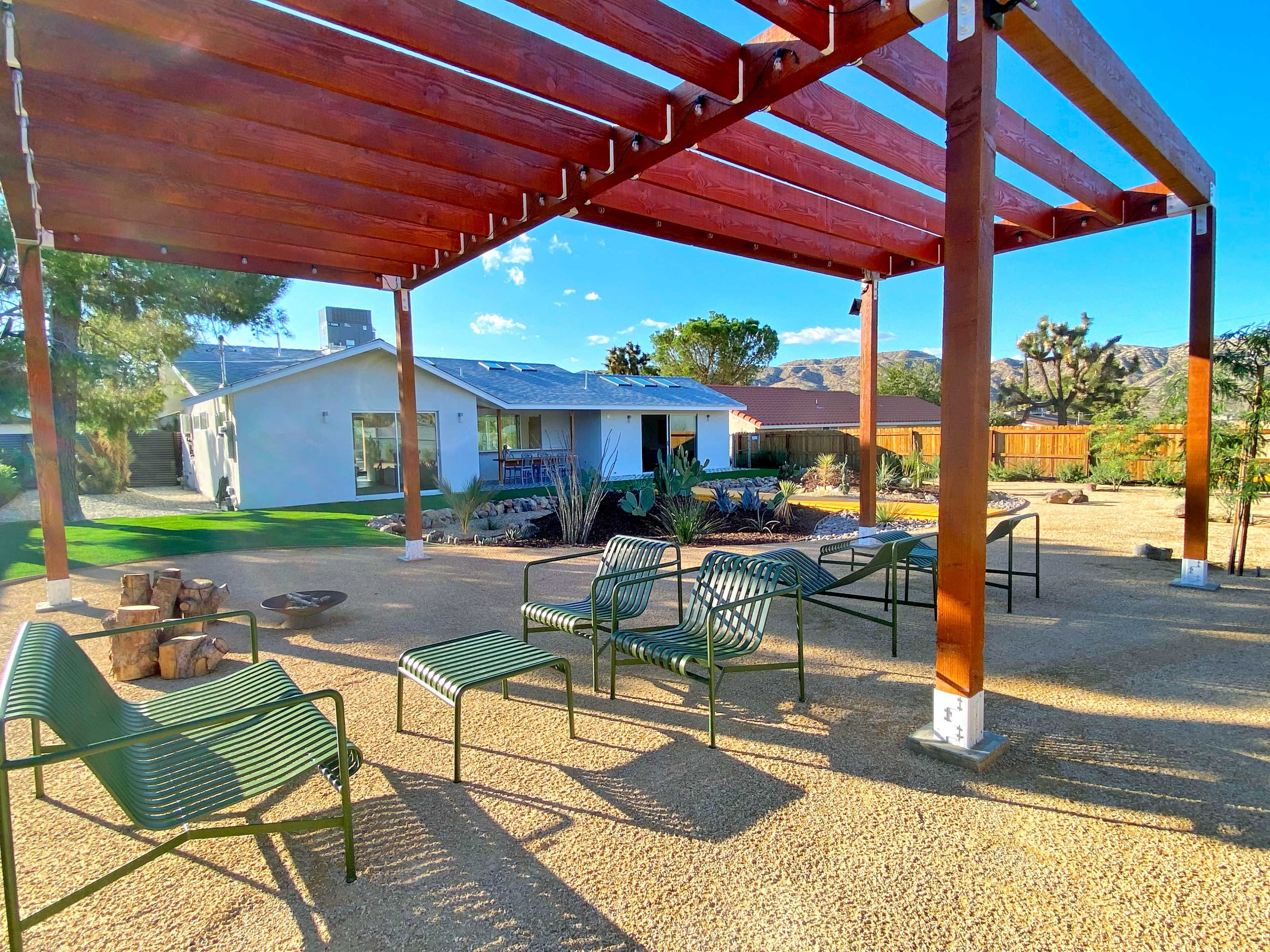 A wooden pergola shaded several green lounge chairs arranged on a gravel patio in a desert landscape with a house in the background.