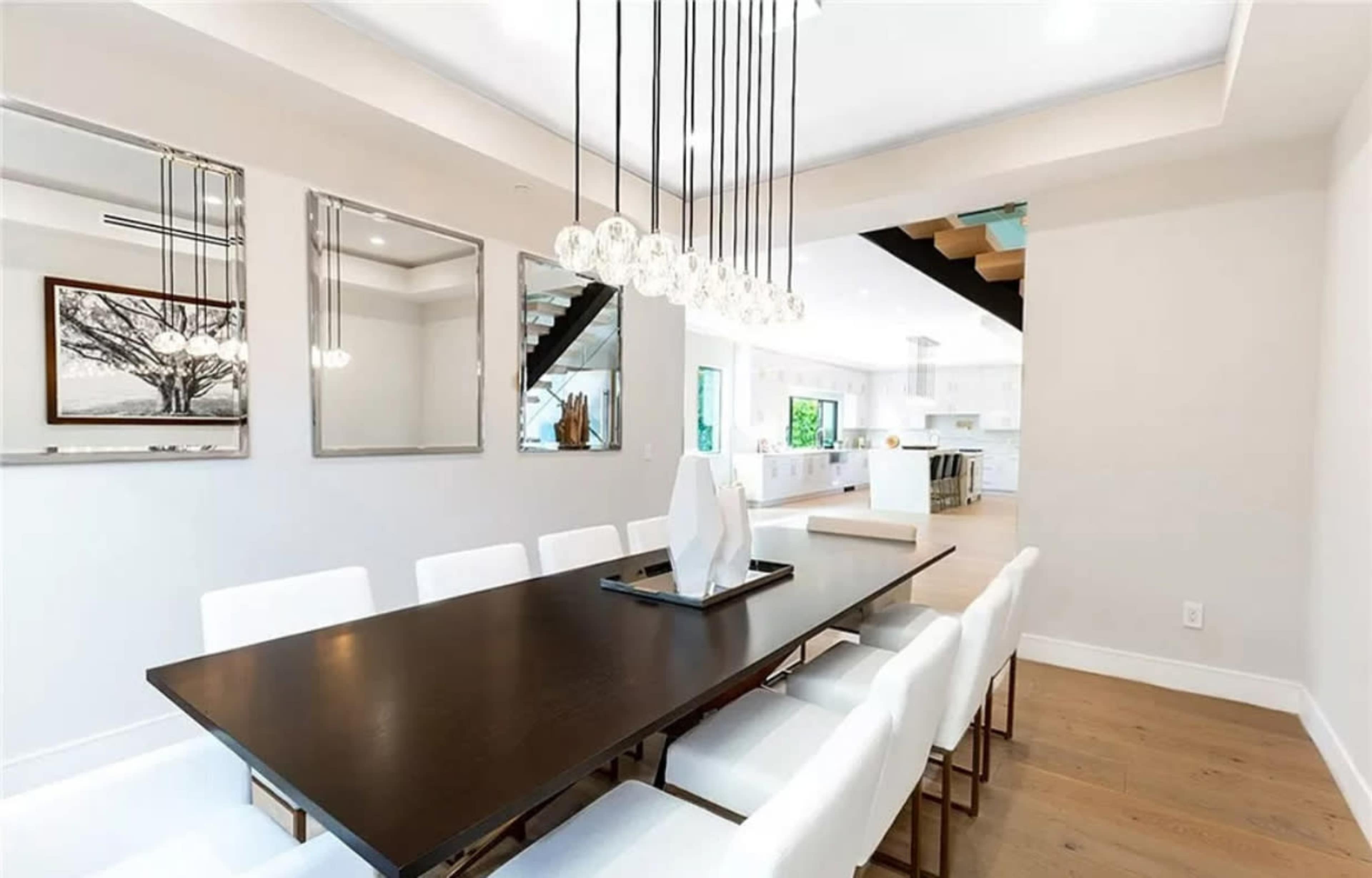 A modern dining room features a long dark wood table surrounded by white chairs, with pendant lighting above and large mirrors on the walls.
