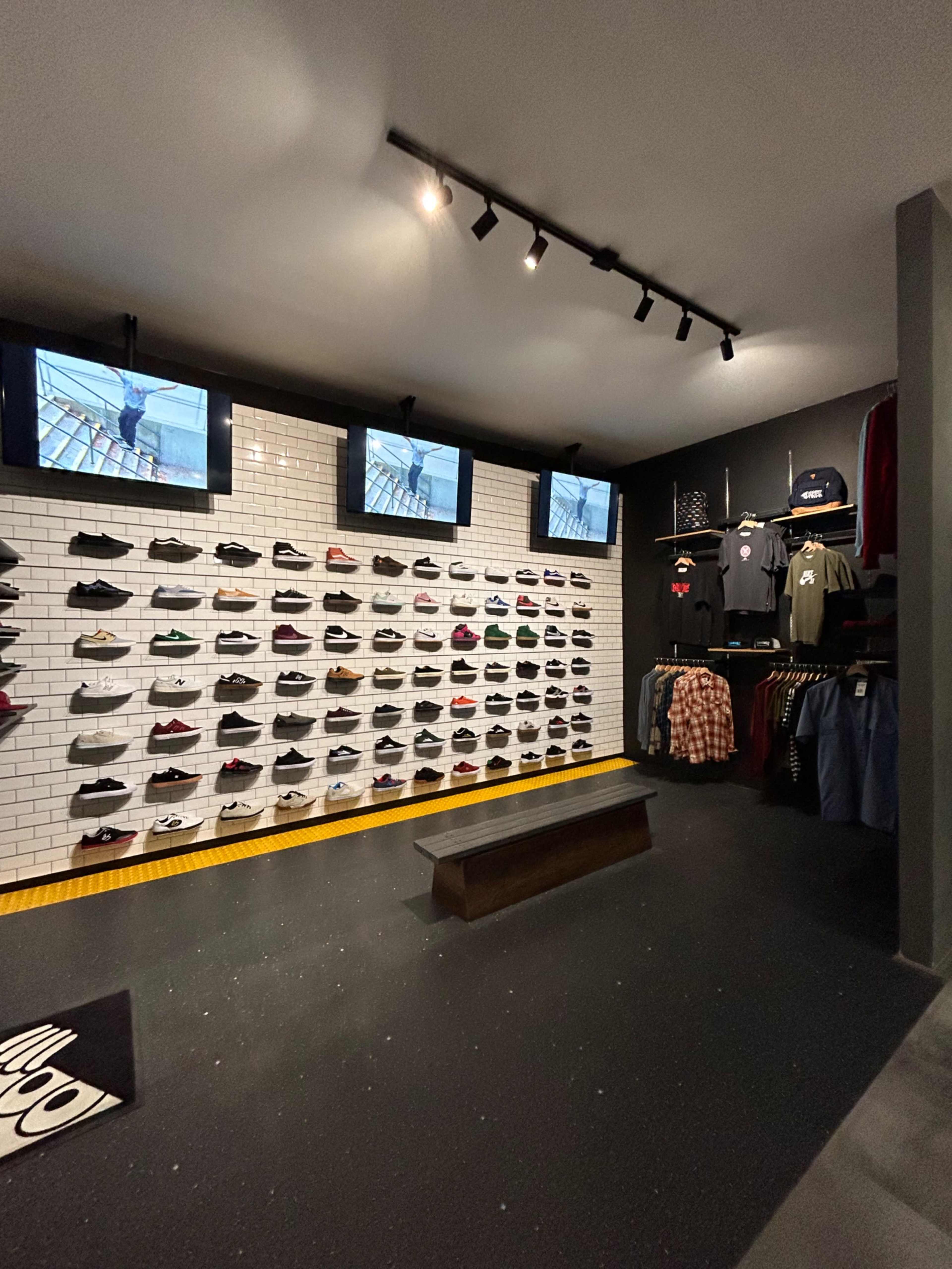 A retail store interior with a shoe display wall, clothing racks, and multiple screens showing videos.