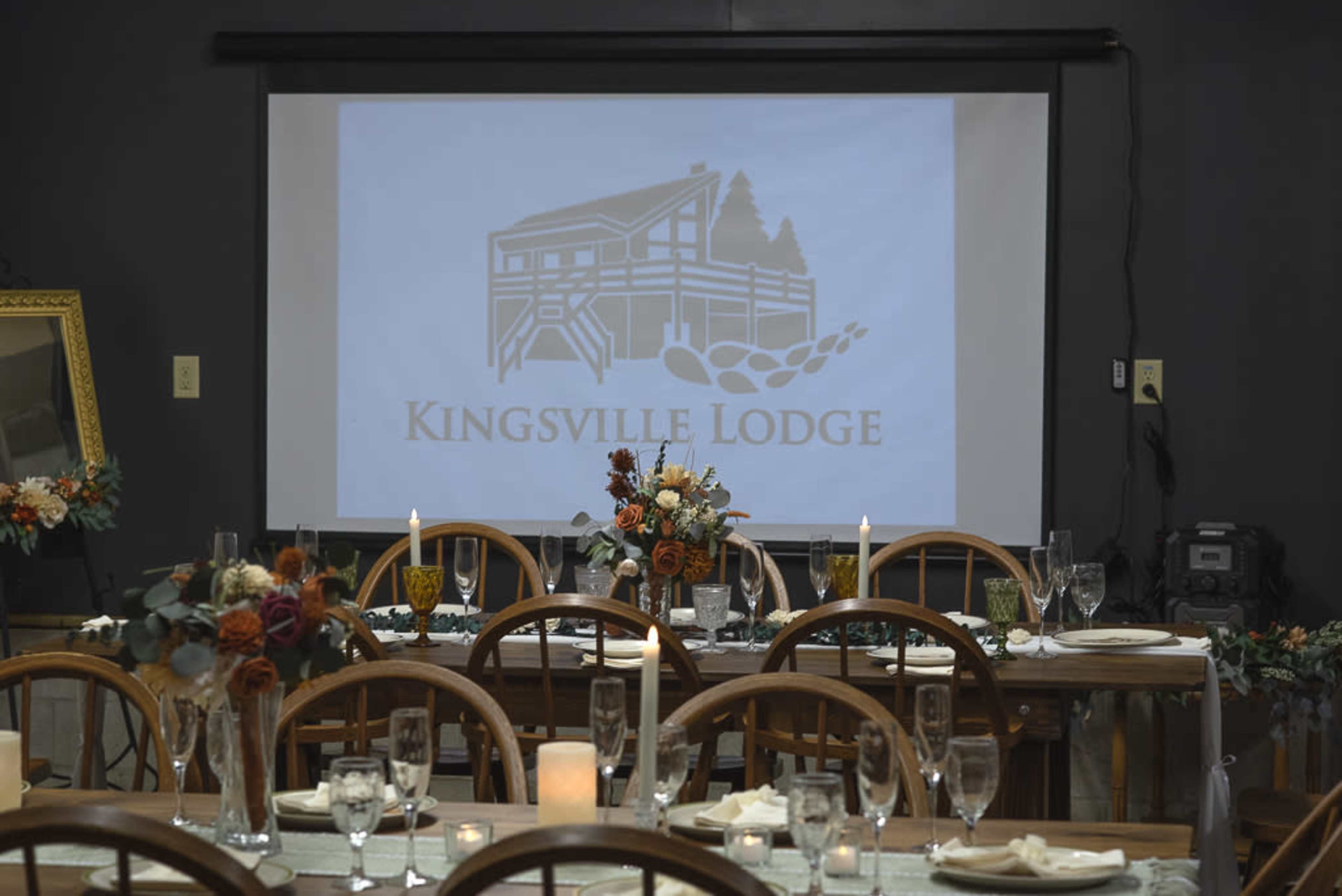 The image shows a dining setup in a lodge with a projector screen displaying the "Kingsville Lodge" logo in the background.