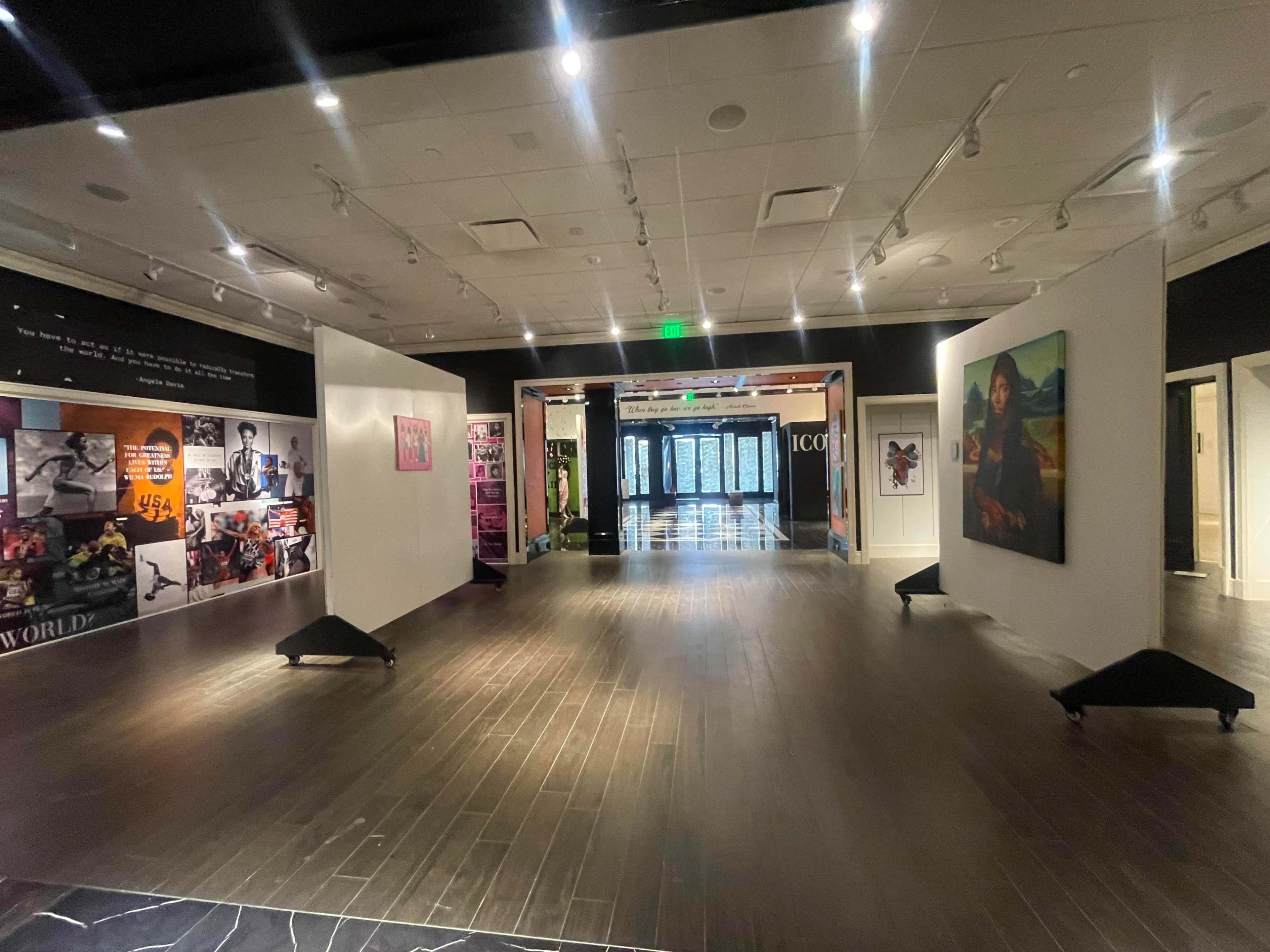 A spacious art gallery entrance featuring white walls with various artworks and photographs displayed, leading to a corridor that opens up to additional exhibition spaces.