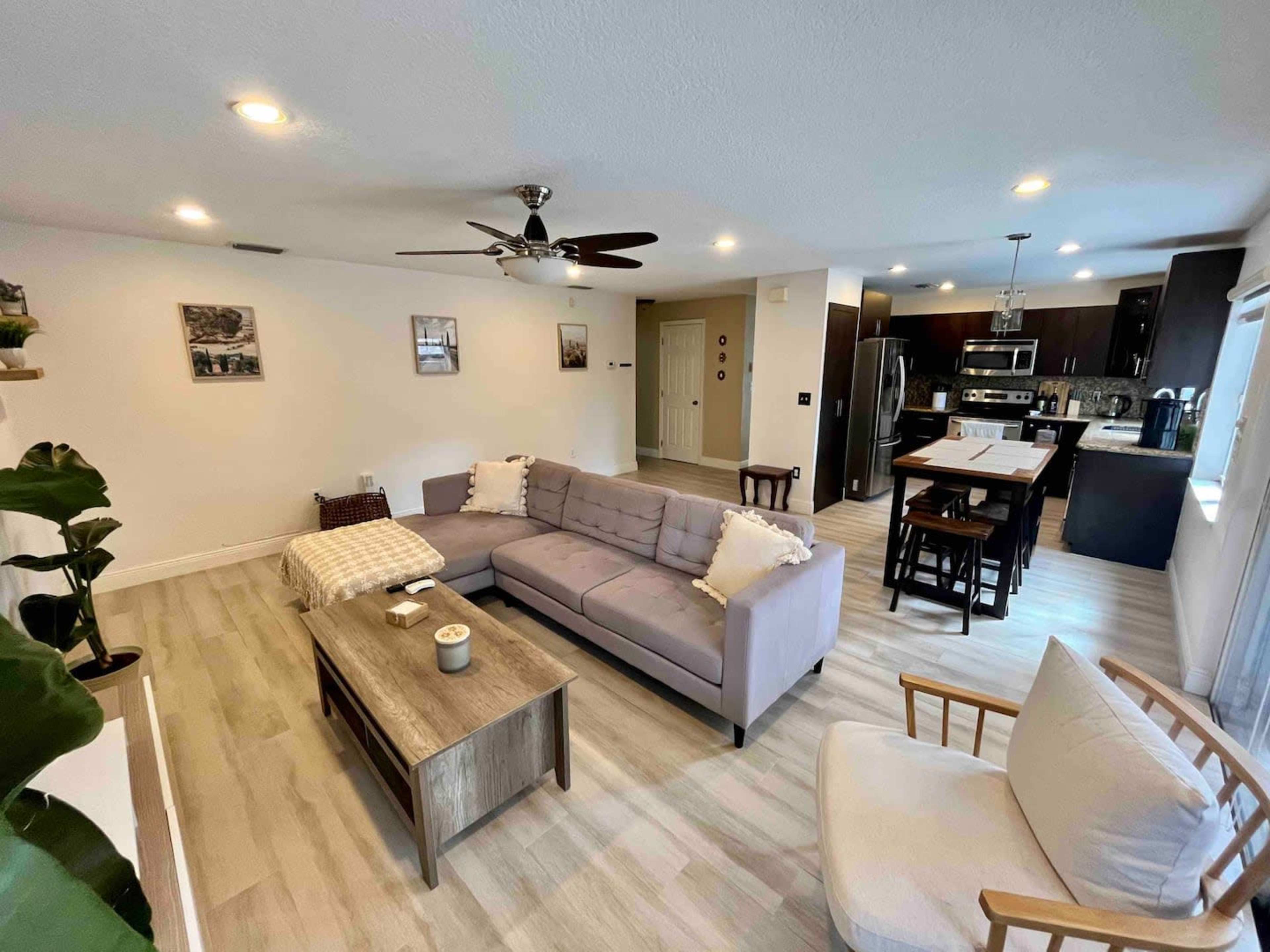 The living area features a light gray sectional sofa, a wooden coffee table, and an open kitchen in the background with dark cabinets and stainless steel appliances.
