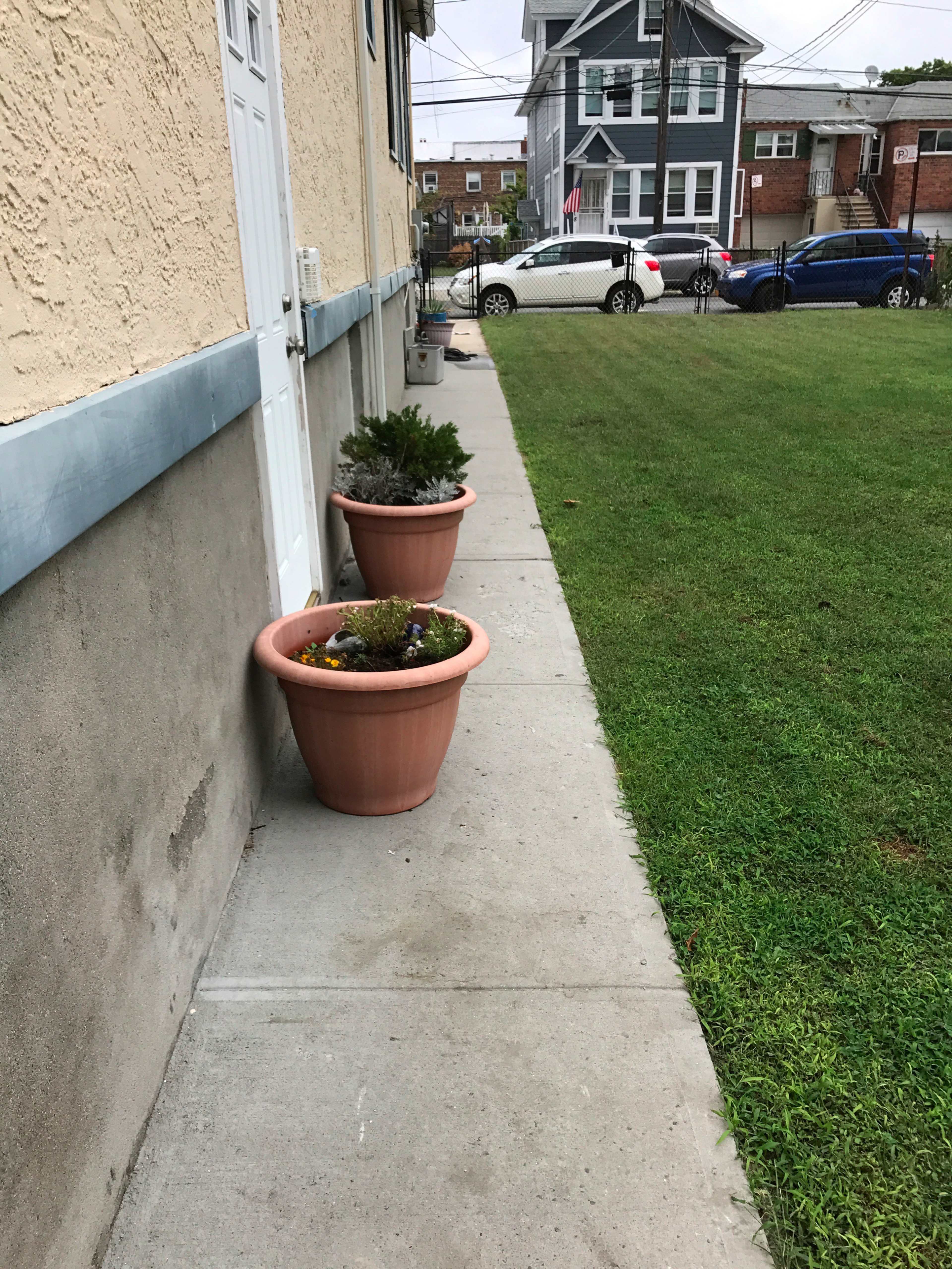 The image shows a concrete sidewalk beside a building, lined with large planters containing plants, leading to a grassy area.