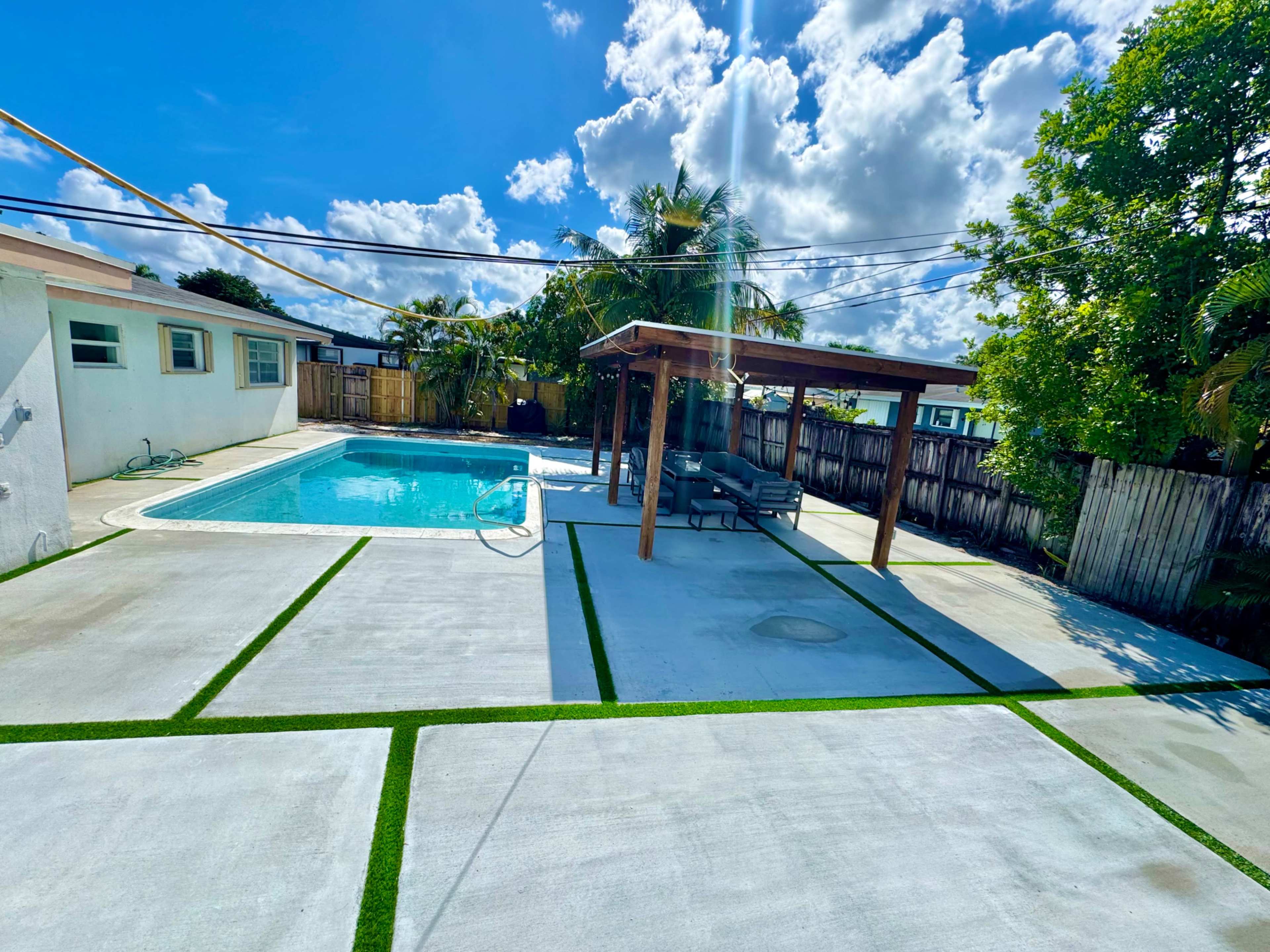 The image shows a backyard with a swimming pool, shaded seating area, and neatly arranged concrete and grass surfaces under a bright blue sky with clouds.