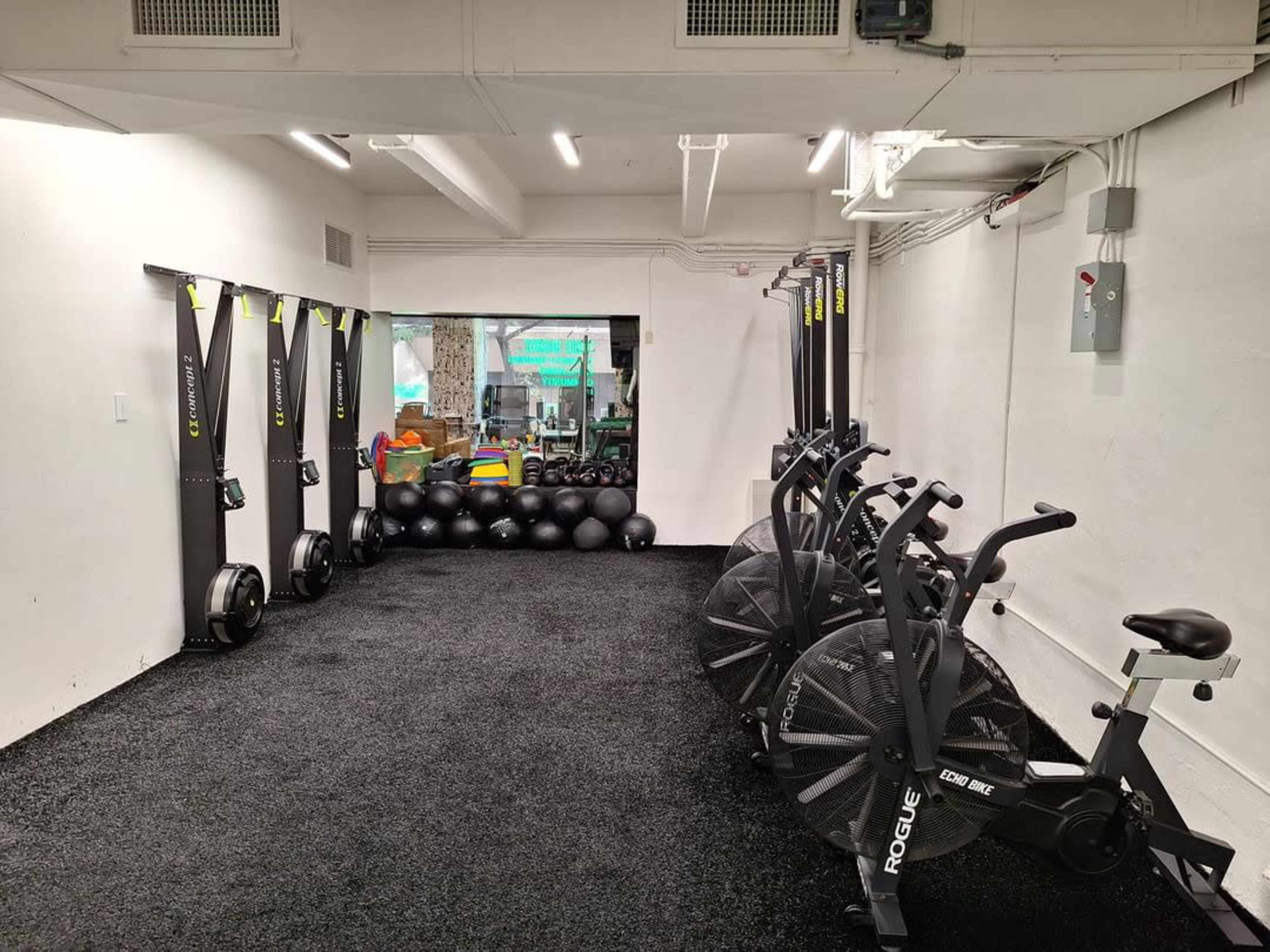 The image shows a fitness room with black exercise equipment, including rowing machines, stationary bikes, and medicine balls arranged against a wall.
