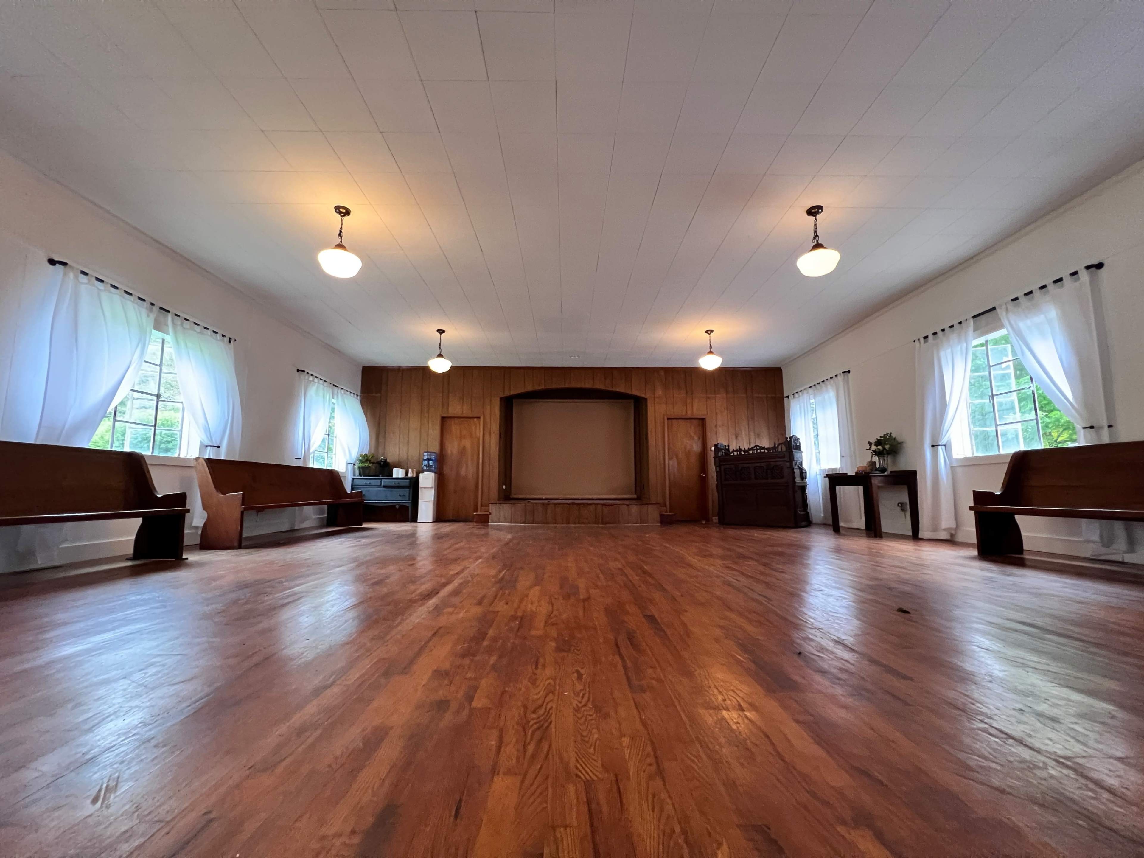 A spacious, empty hall with wooden floors, benches along the sides, and large windows with white curtains.