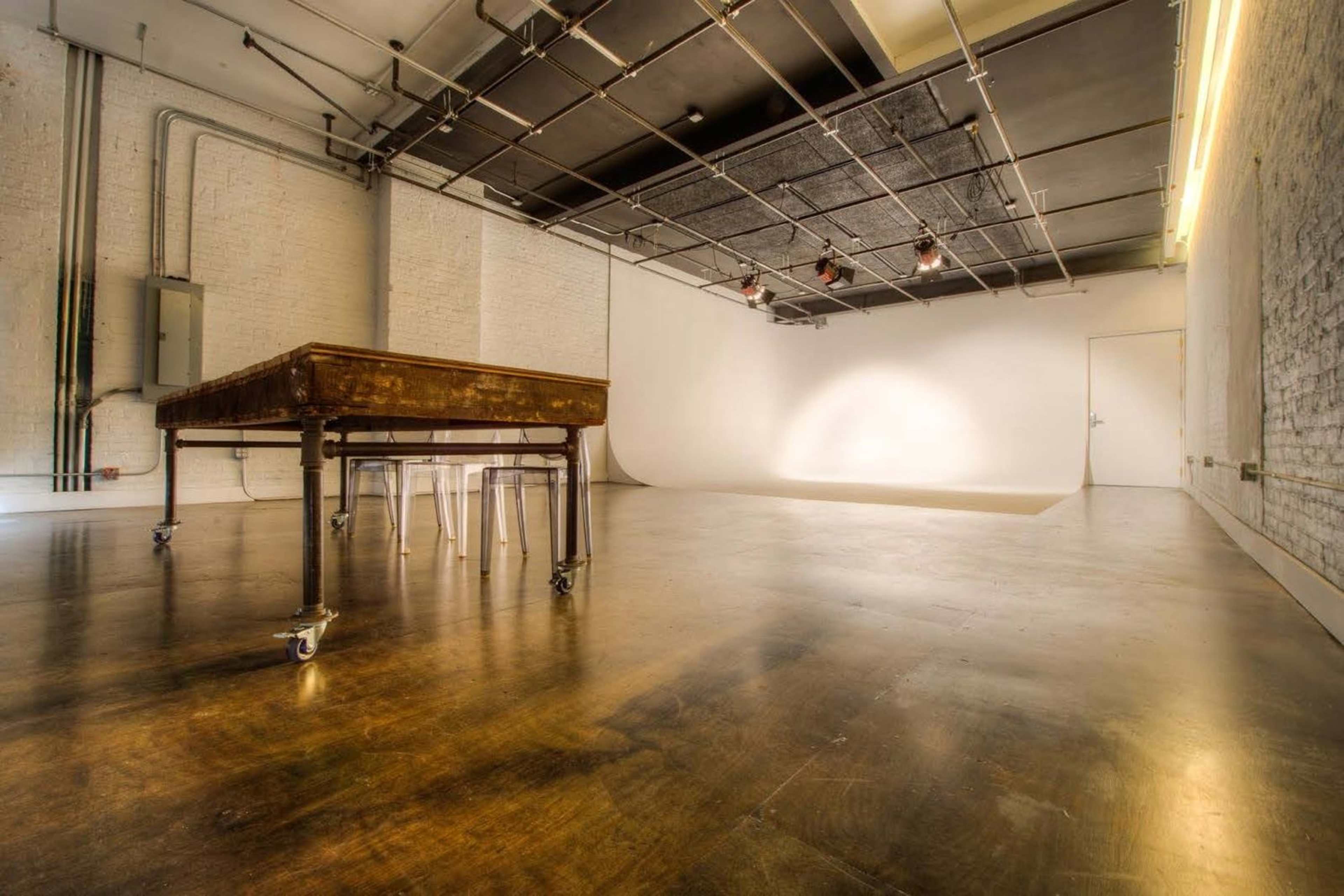 A spacious, minimalist studio features a large table on wheels and clear chairs, with a blank backdrop and overhead lighting.