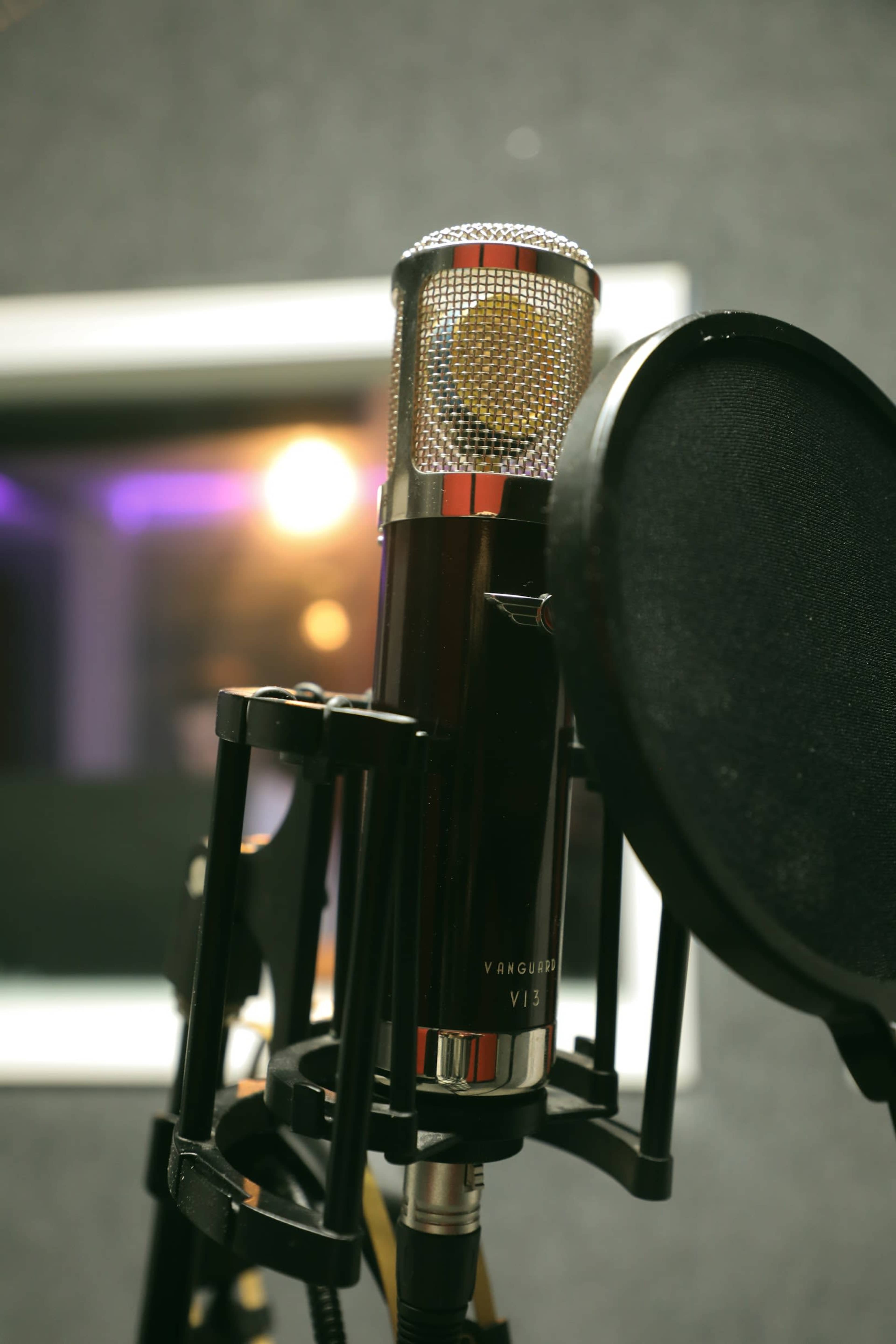 A close-up of a microphone mounted on a stand with a pop filter in front of it, set against a blurred backdrop of a recording studio.