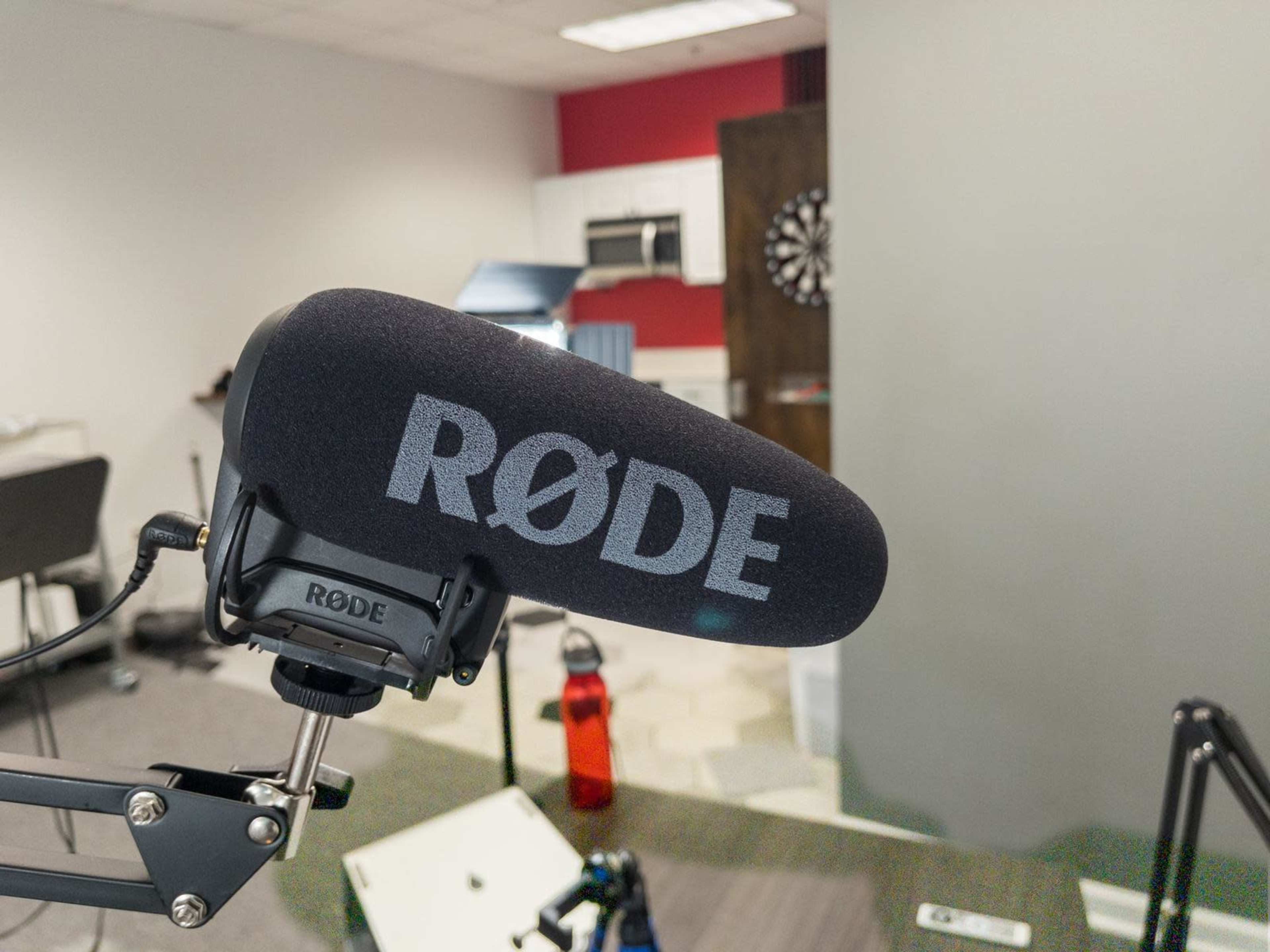 A close-up of a Rode microphone attached to a boom arm, with a blurred background featuring a kitchen and a dartboard.