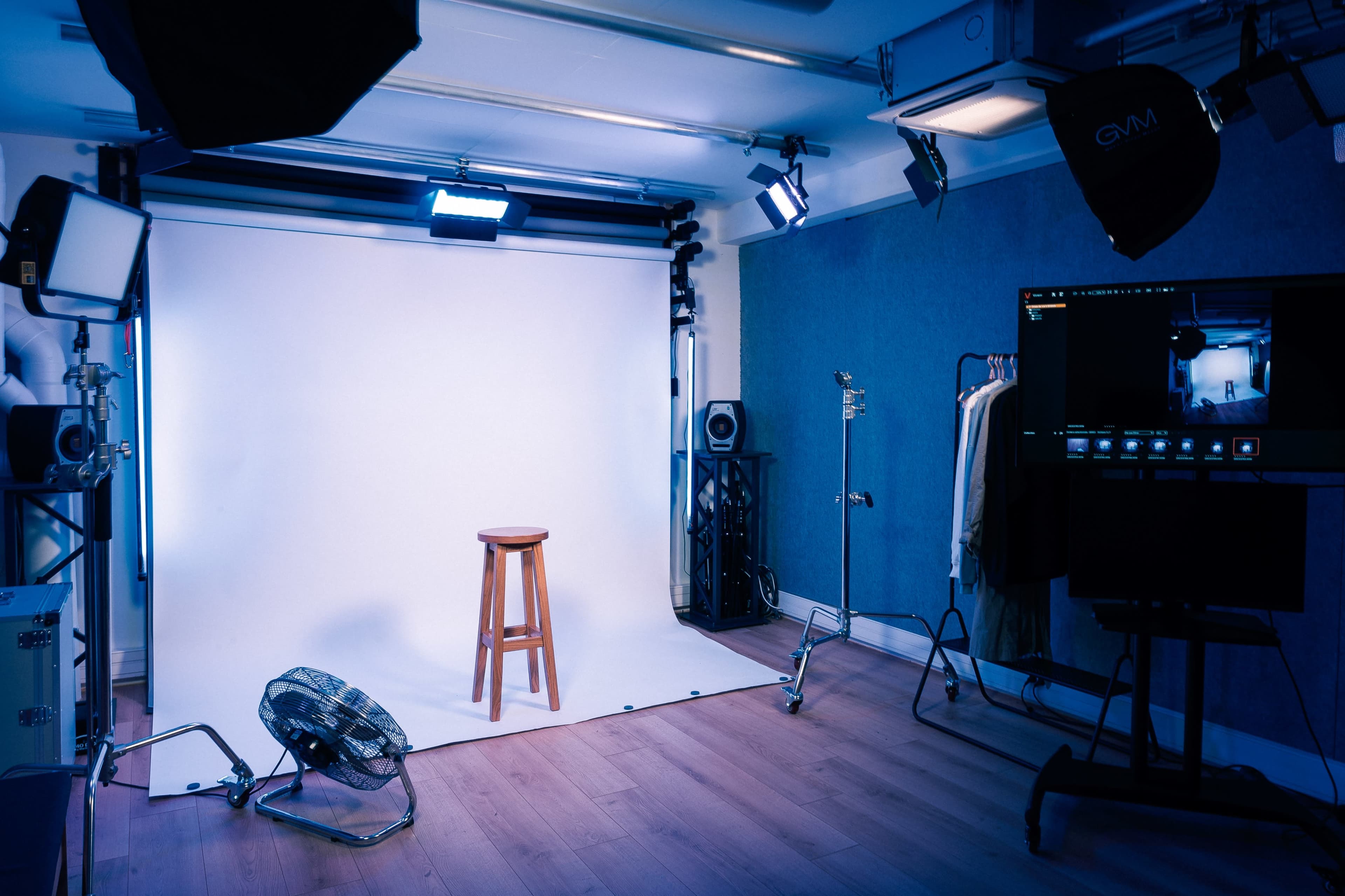 A photography studio with a wooden stool positioned in front of a white backdrop, surrounded by studio lights and various equipment.