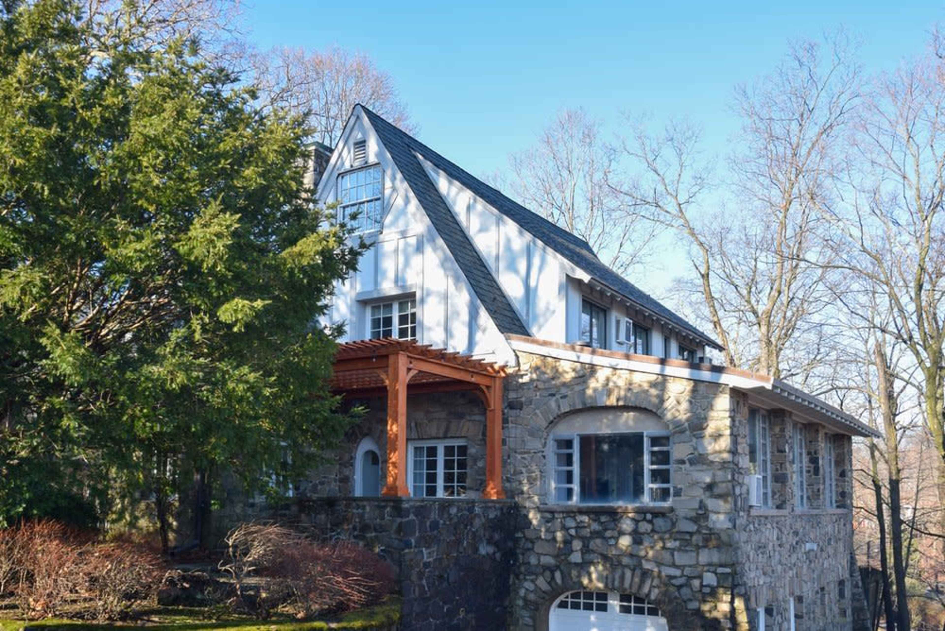 The image shows a two-story house with a stone base, wooden porch, and a sloped roof, situated among bare trees.