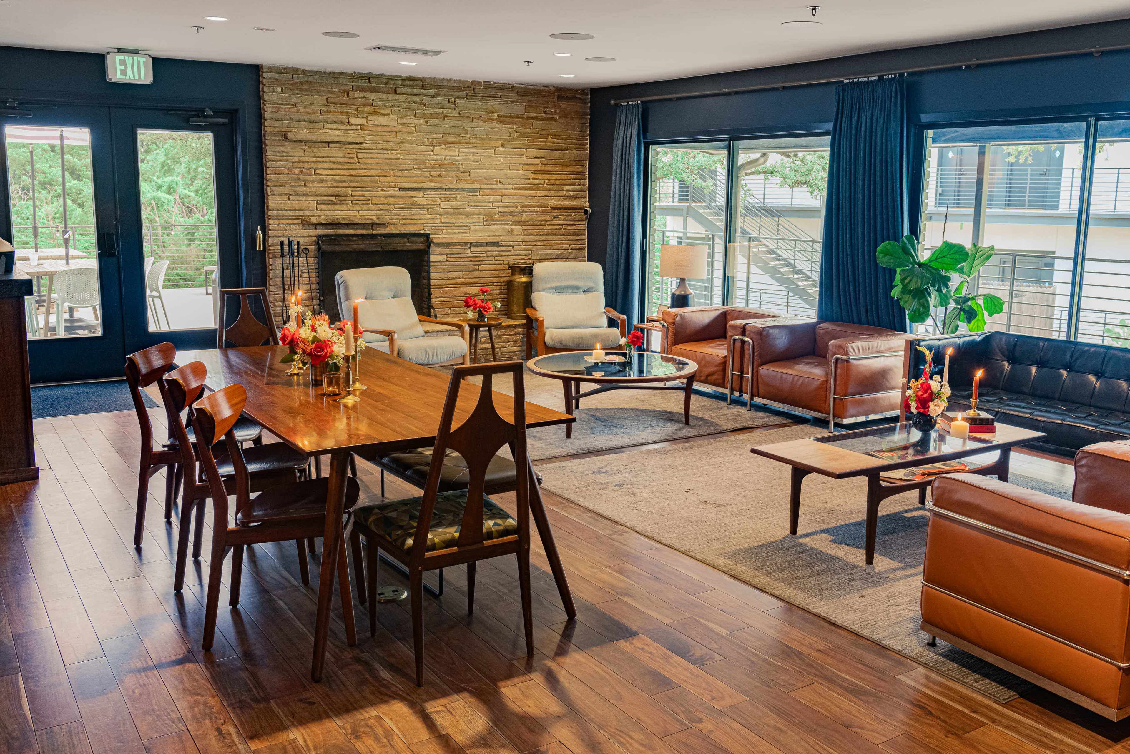 The image shows a spacious lounge area featuring a wooden dining table, leather sofas, and a stone wall with large windows providing natural light.