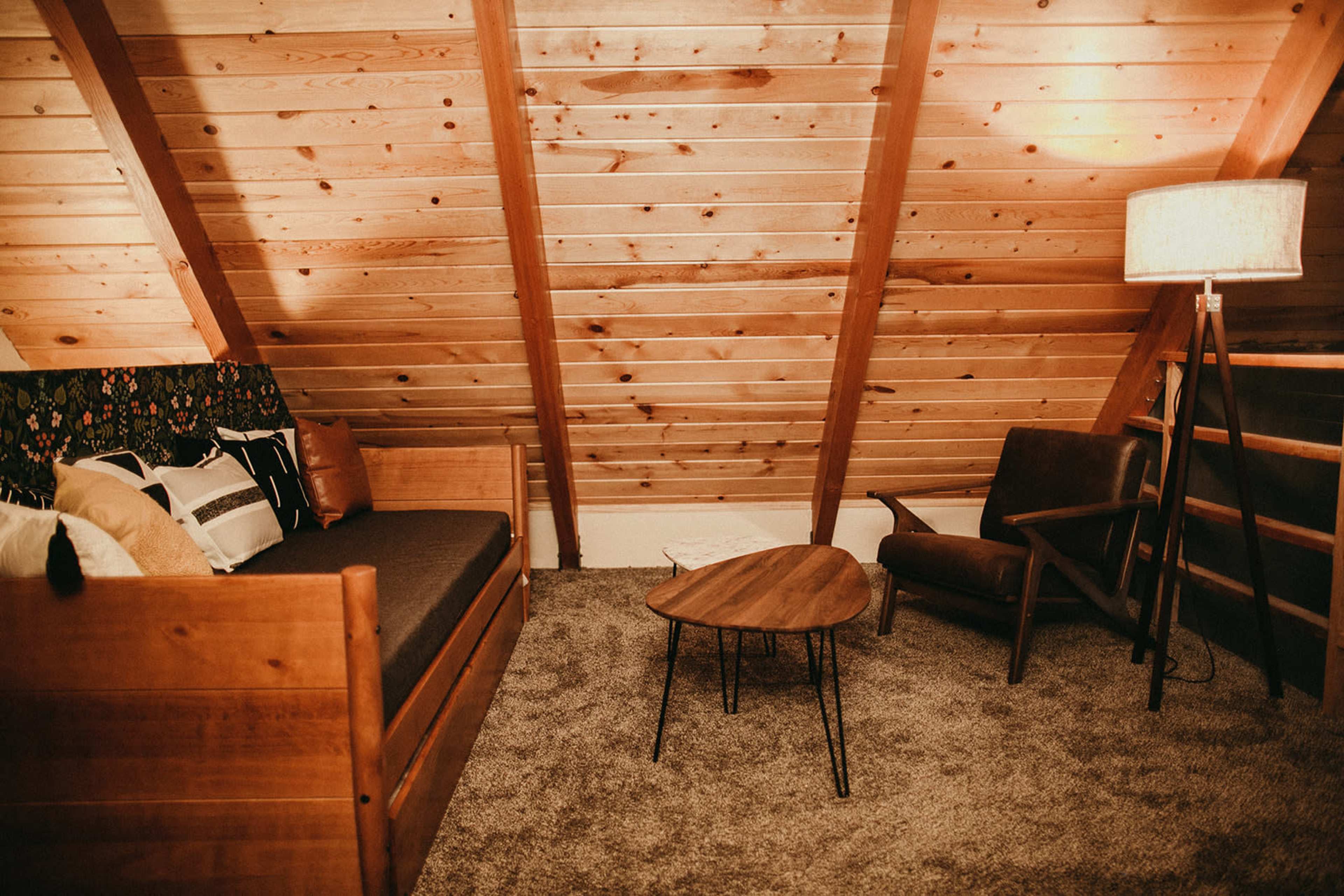 A cozy loft space with wooden beams, a sofa, a chair, and a round coffee table on a carpeted floor.
