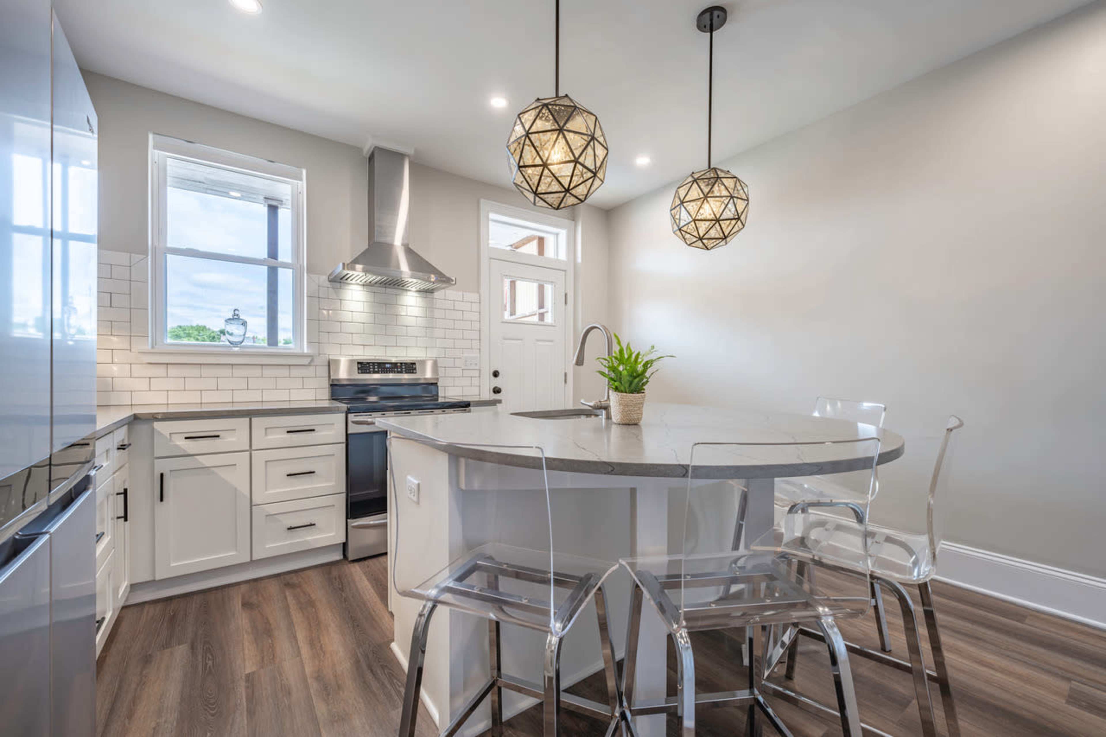 A modern kitchen features a round island with transparent stools, pendant lighting, stainless steel appliances, and a window above the sink.