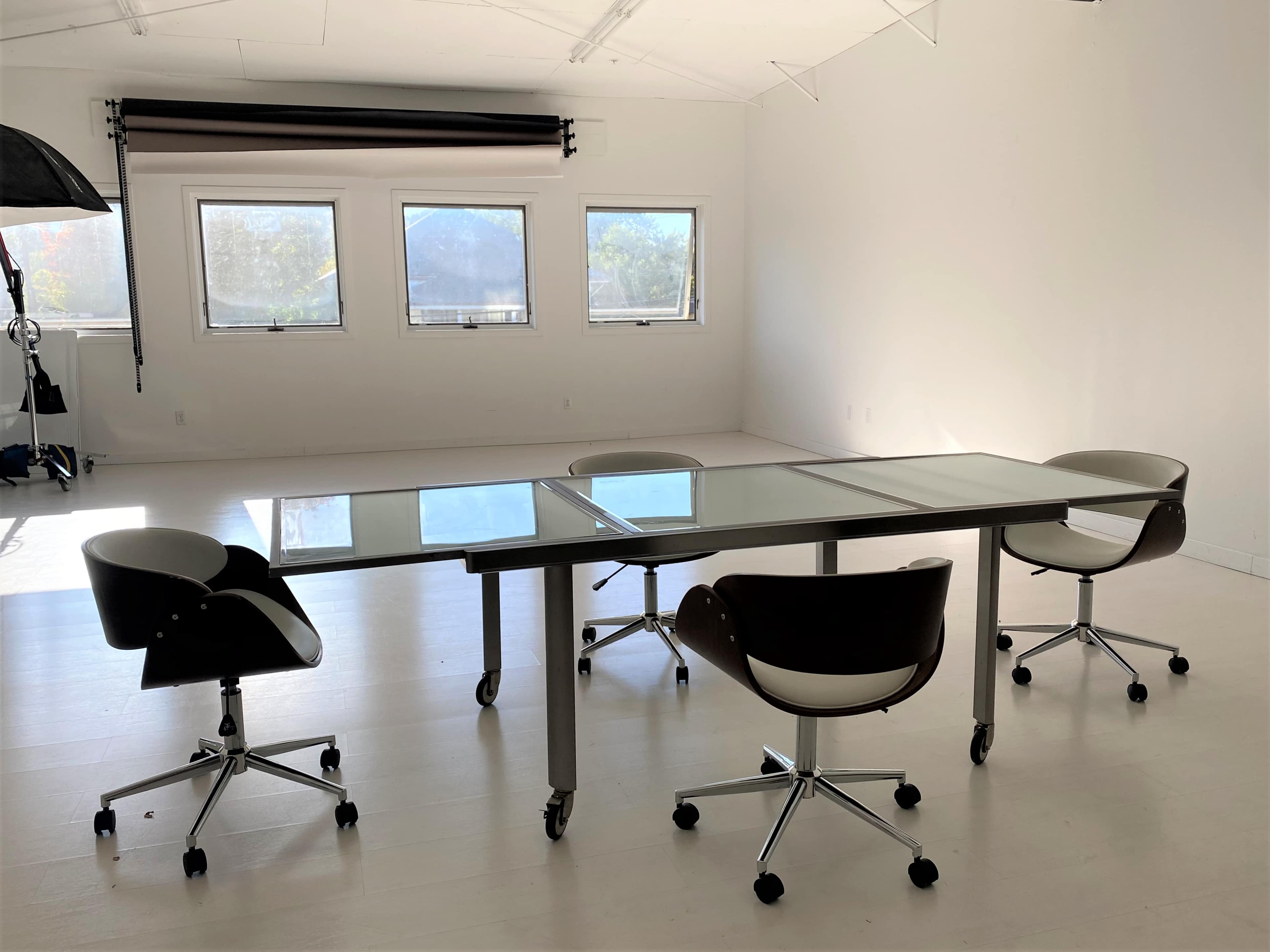 A modern meeting room features a glass-top table surrounded by four black and white rolling chairs, with large windows letting in natural light.
