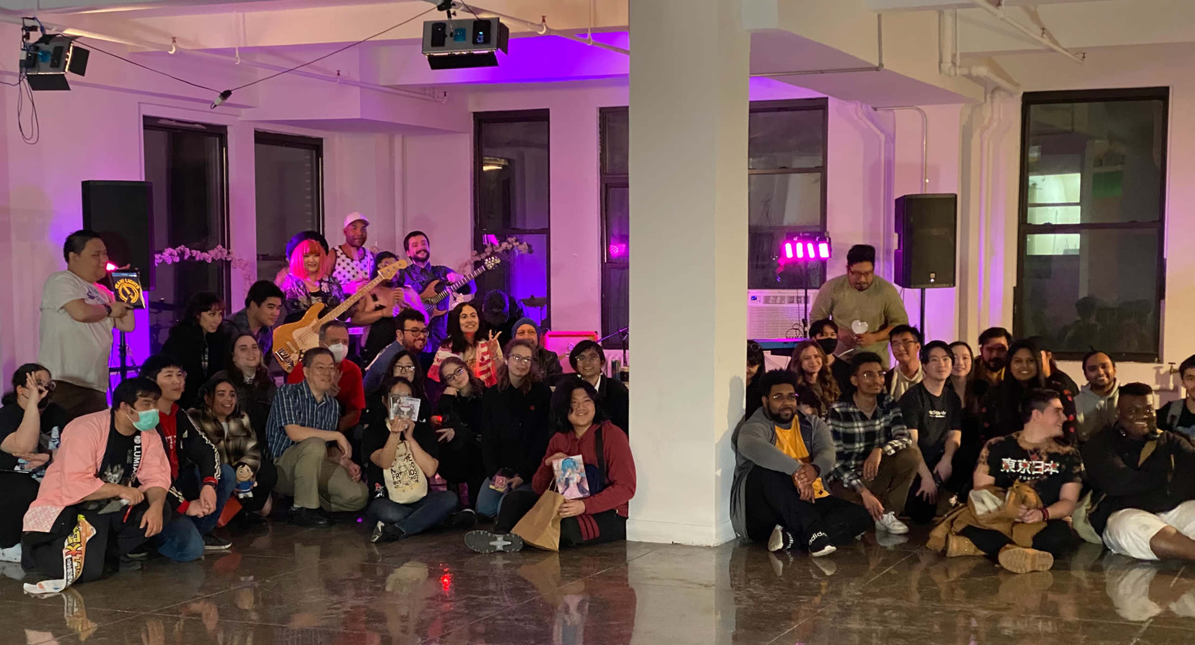 A large group of people poses together in a brightly lit room with a stage set up for a musical performance in the background.