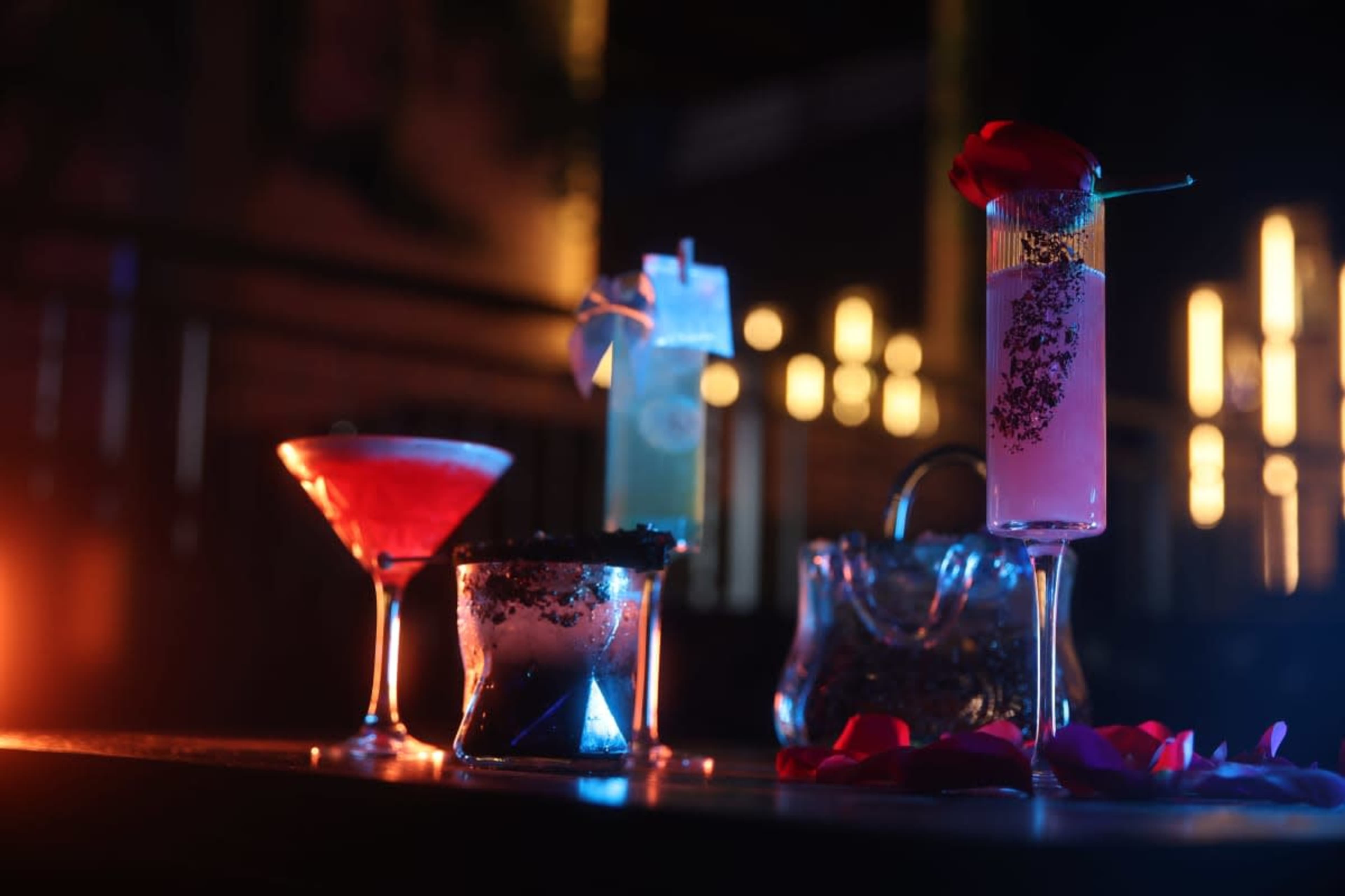 The image displays a variety of colorful cocktails elegantly arranged on a bar, illuminated by dim lighting and surrounded by rose petals.