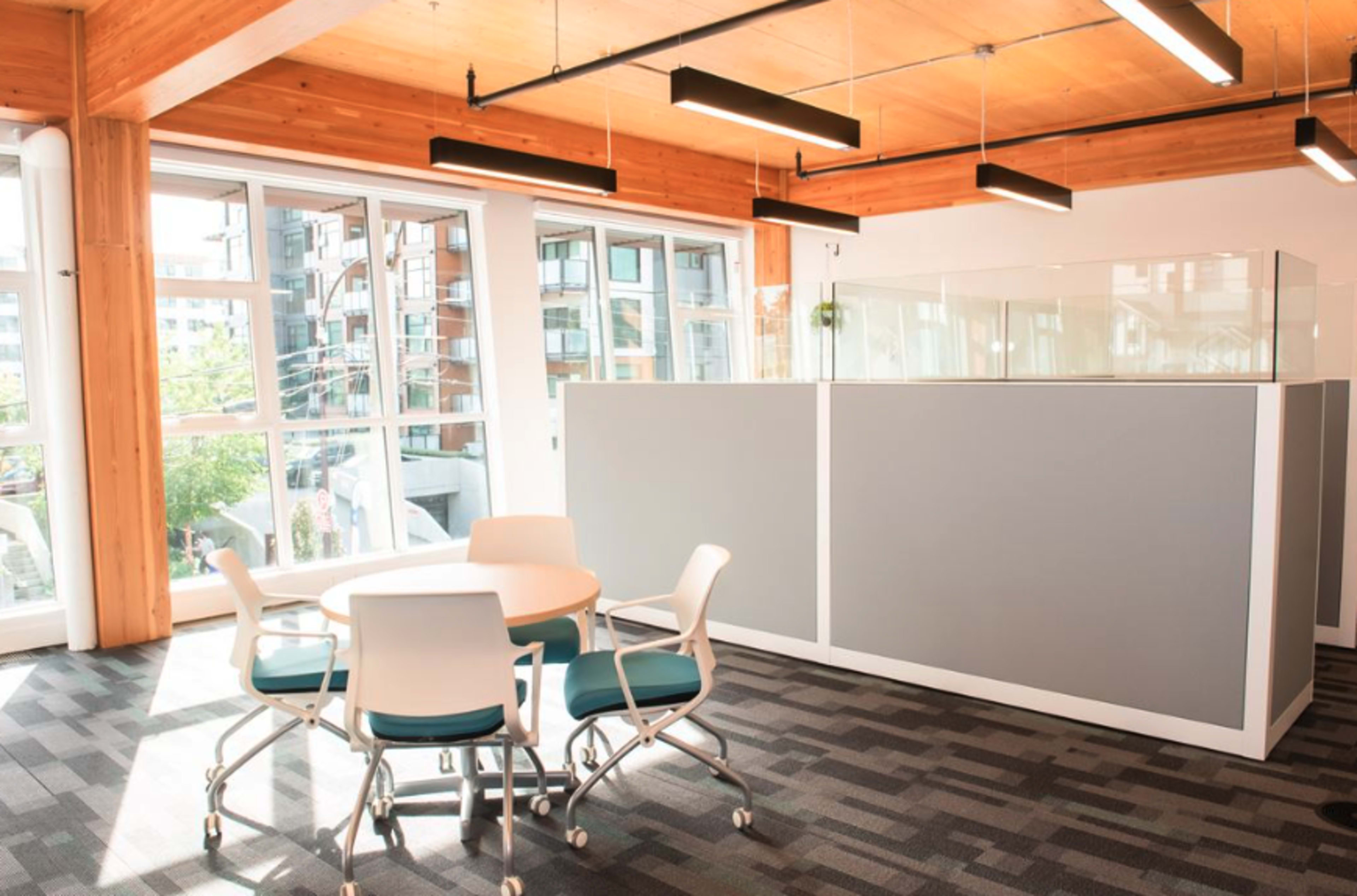 A minimally furnished office space features a round table with four chairs near a glass-enclosed area and large windows.