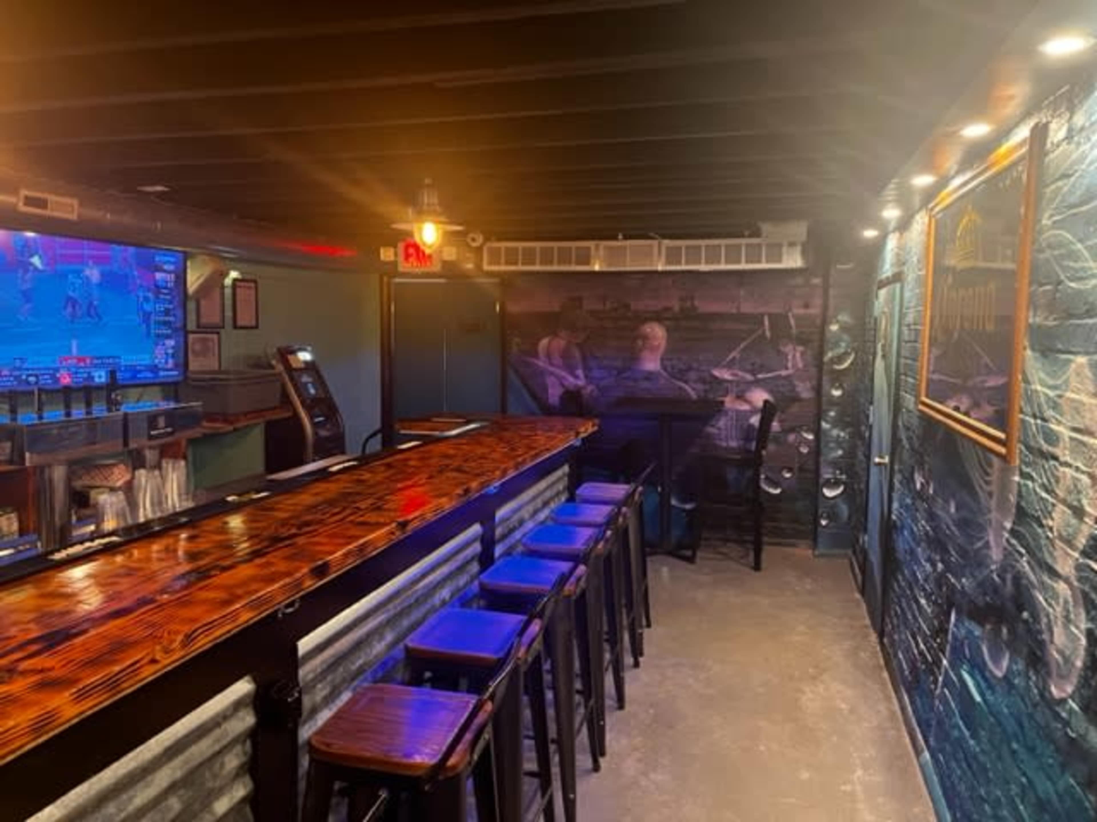 The interior of a bar featuring a long wooden counter, high stools, a video screen displaying a sports game, and a mural on the wall.