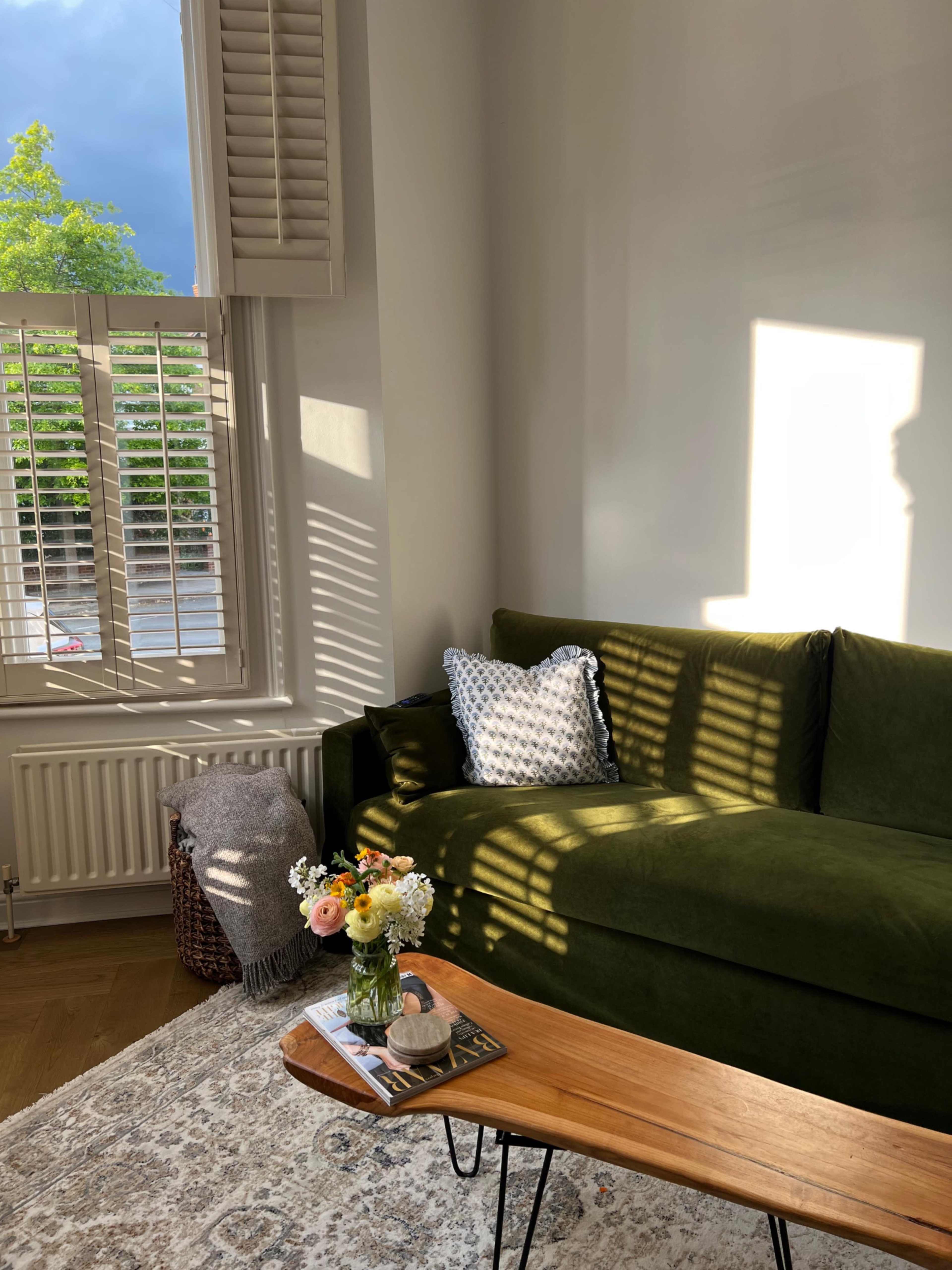 A green sofa with patterned cushions sits beside a wooden coffee table adorned with a vase of flowers and a decorative object, illuminated by sunlight streaming through the window.