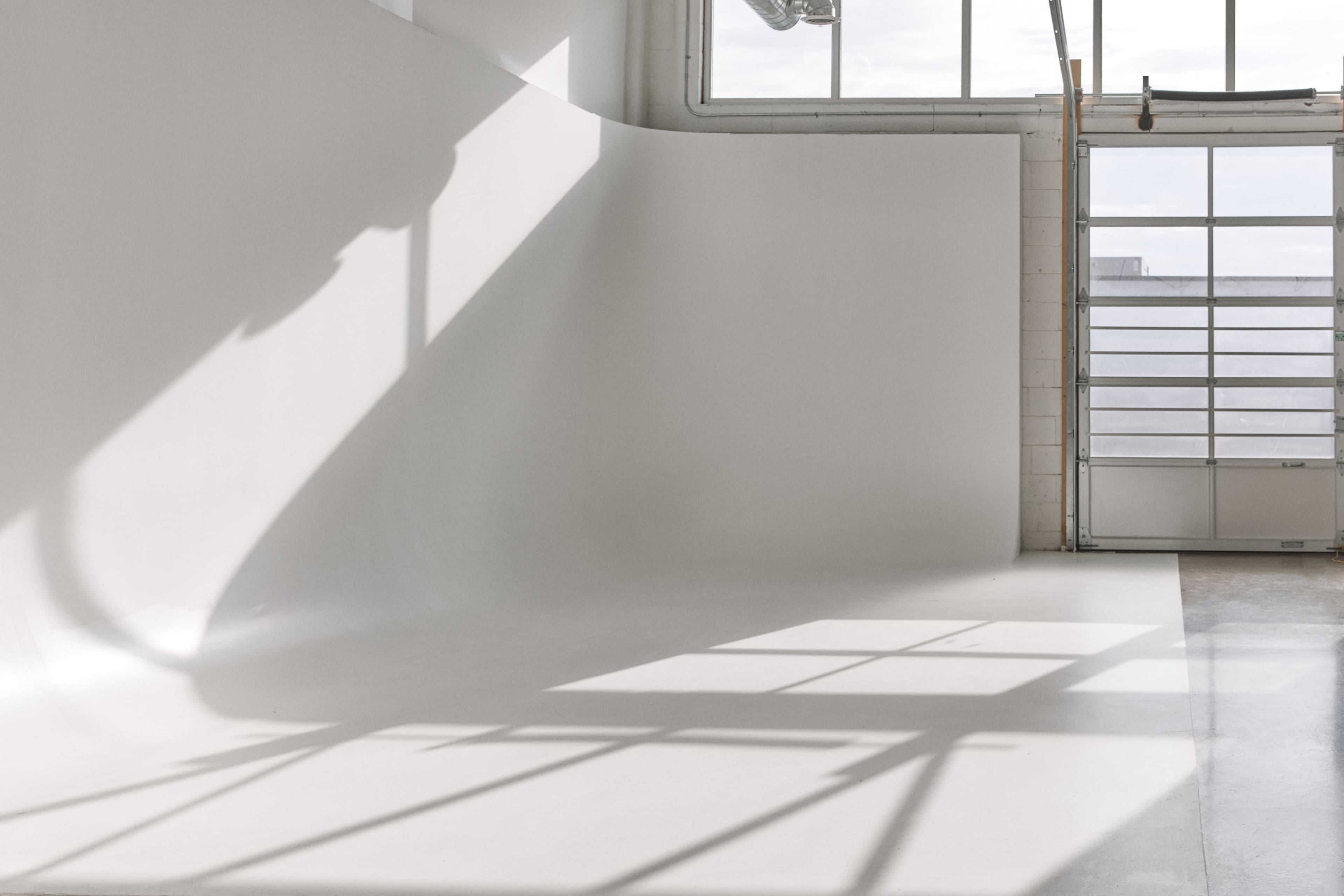 A bright studio space with a curved white wall and large windows casting shadows on the floor.