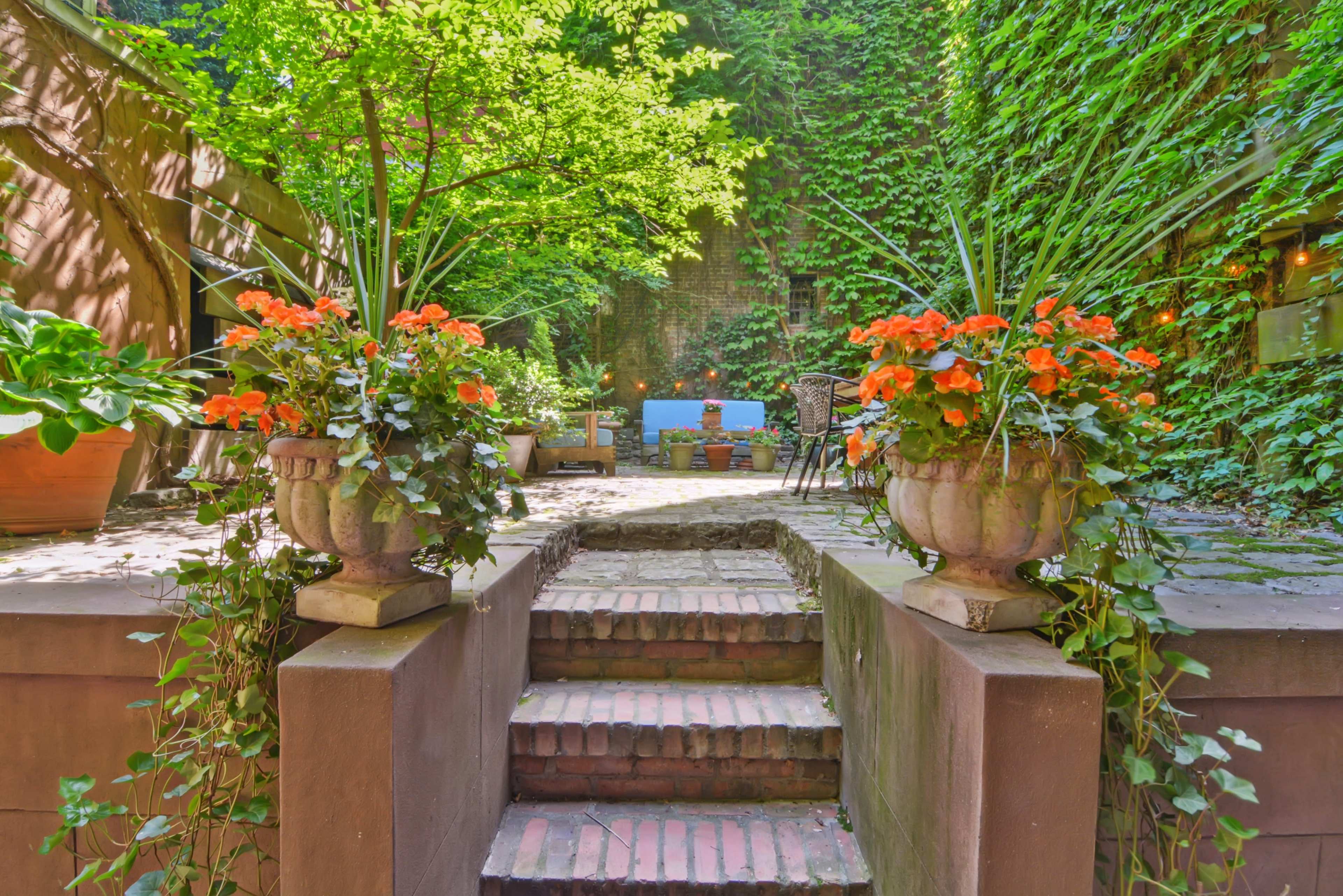 The scene features a brick pathway leading up stone steps, flanked by flower pots with orange flowers, and surrounded by a lush, green garden with ivy-covered walls.