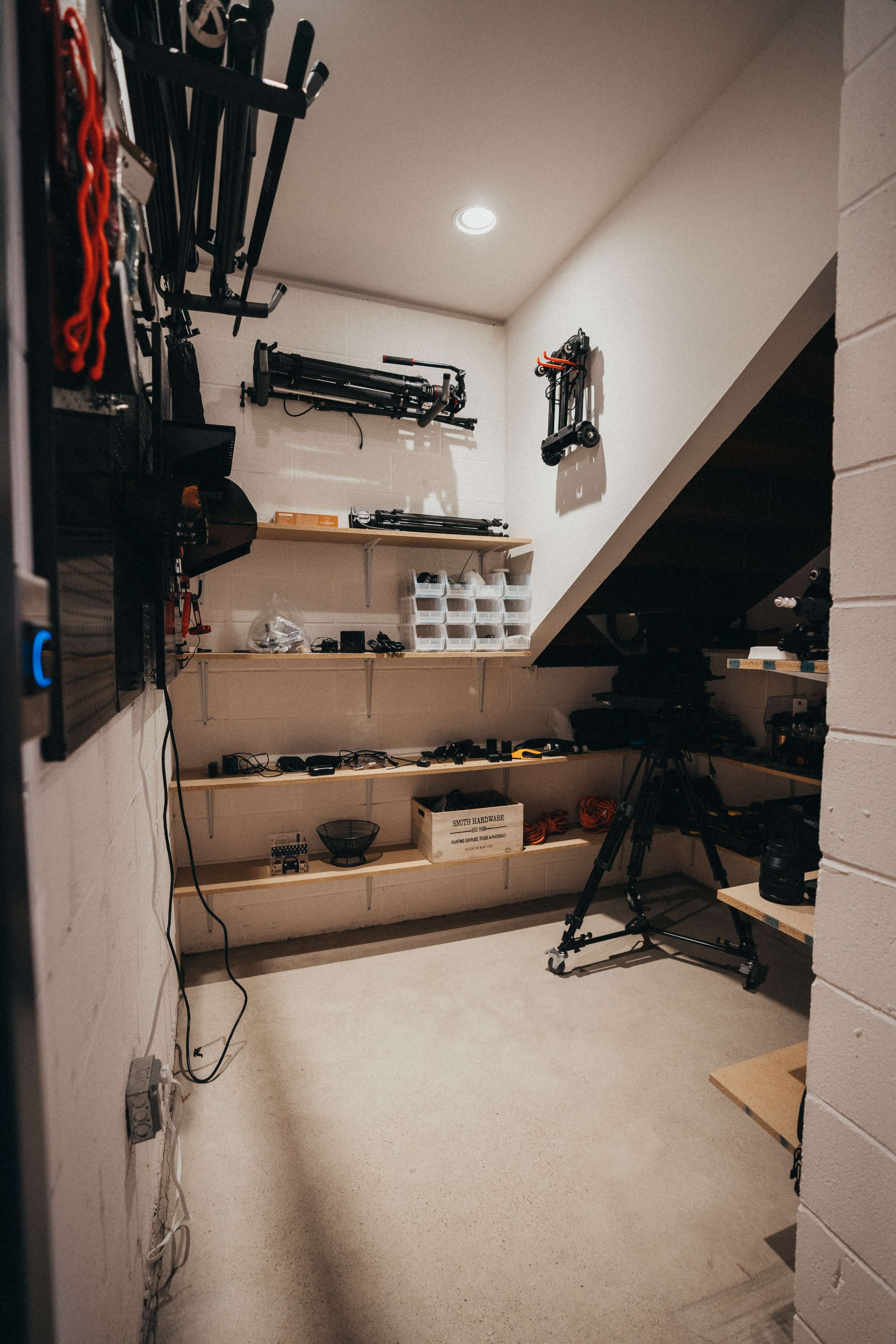 A storage room under a staircase, filled with shelves holding various camera equipment and accessories.