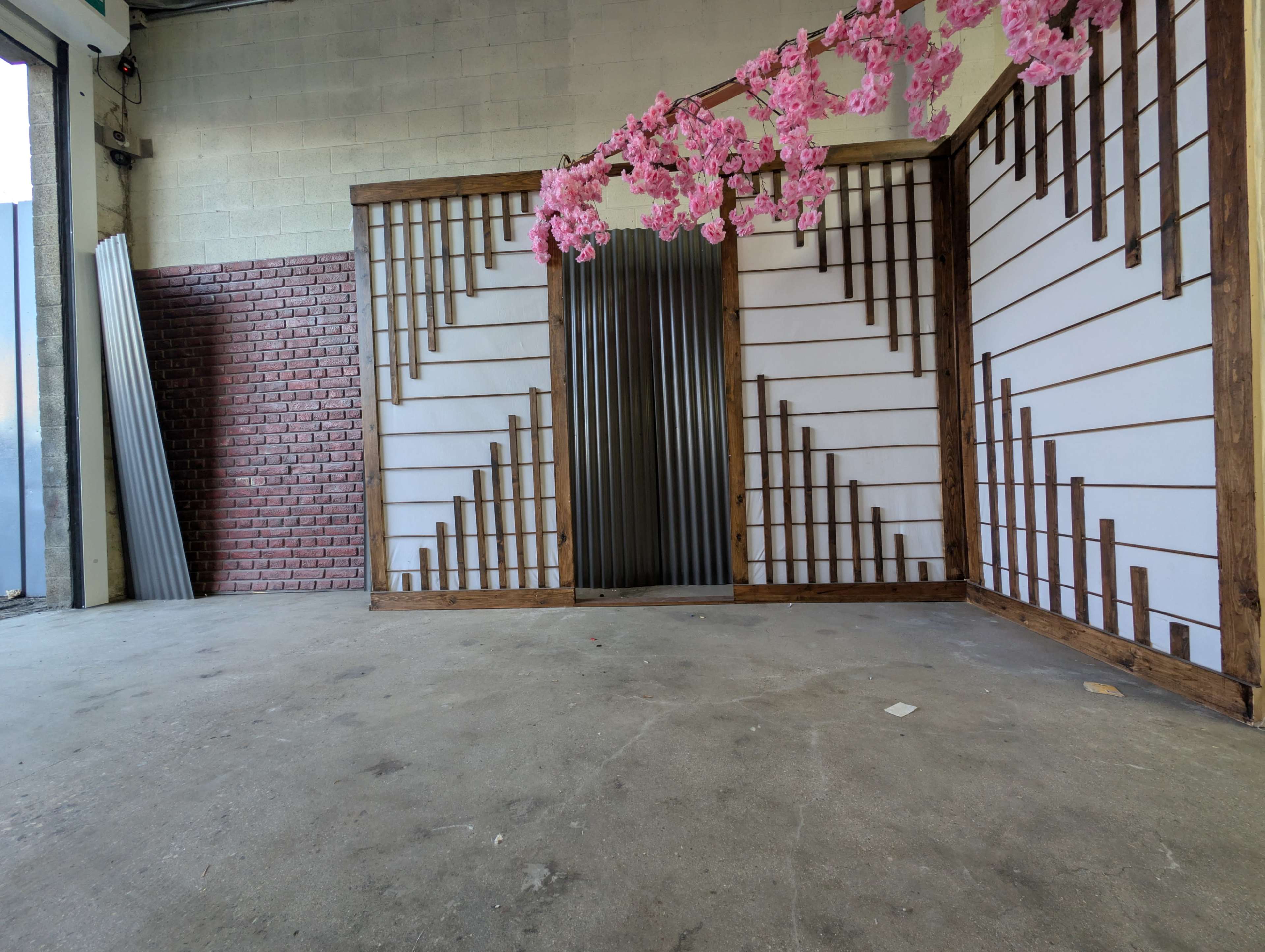 The image shows an empty room with a patterned wooden wall, a metal door in the center, and artificial cherry blossom branches hanging overhead.