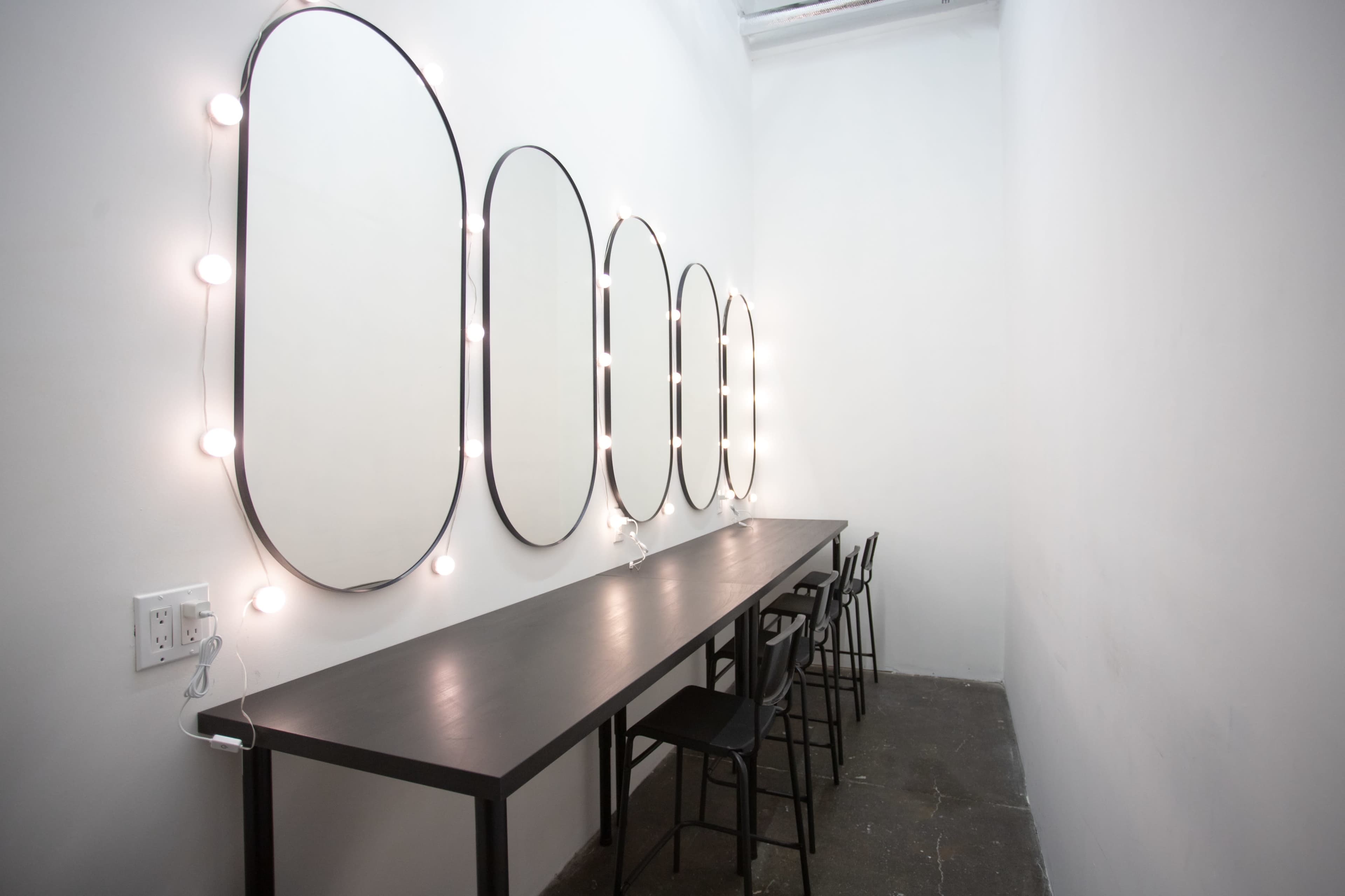 A narrow dressing room with a long table and several oval mirrors lit by bulbs along the walls.