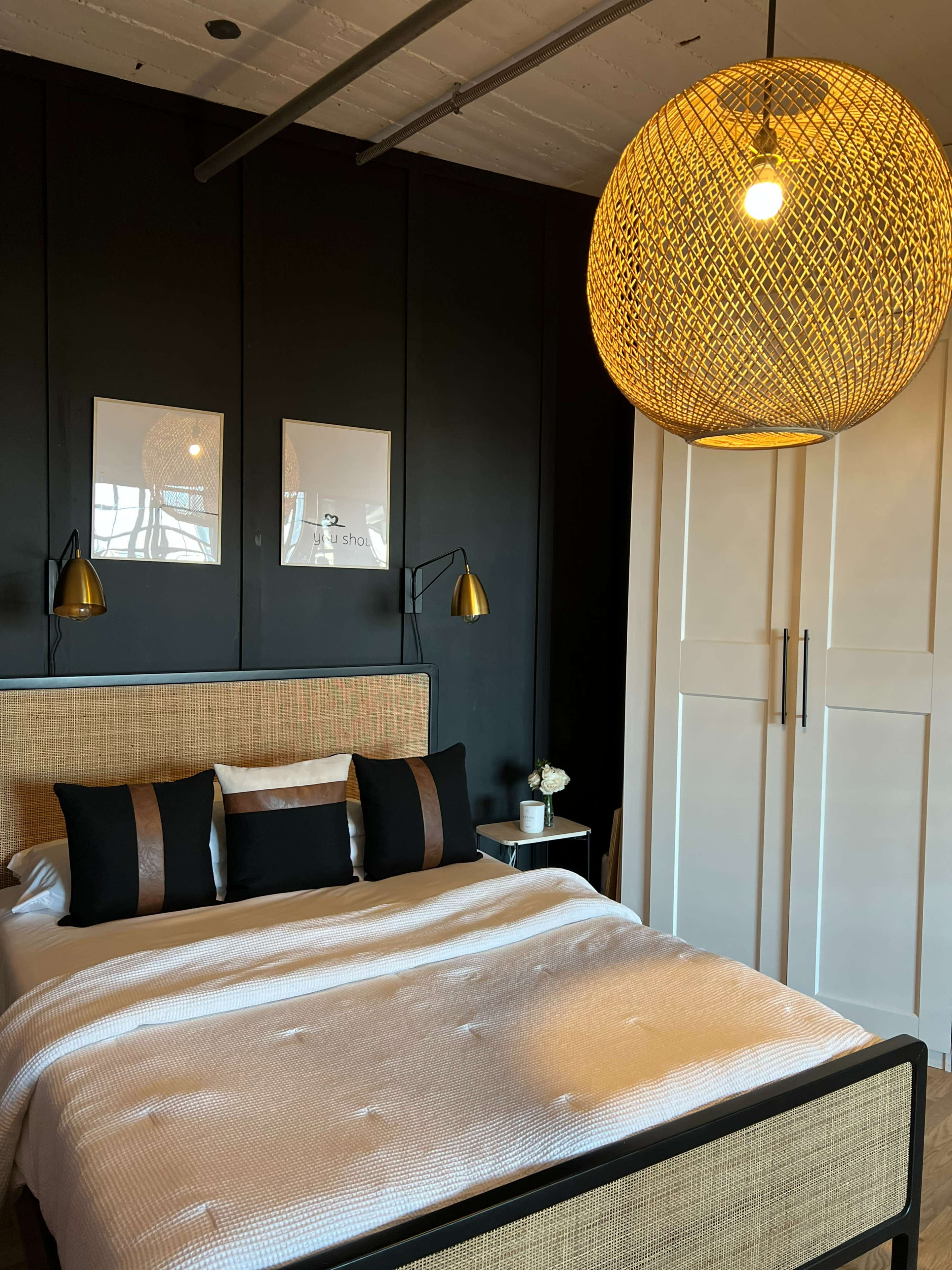 A modern bedroom featuring a bed with a white quilted coverlet, black pillows, a woven pendant light, and a black accent wall with two framed artworks.