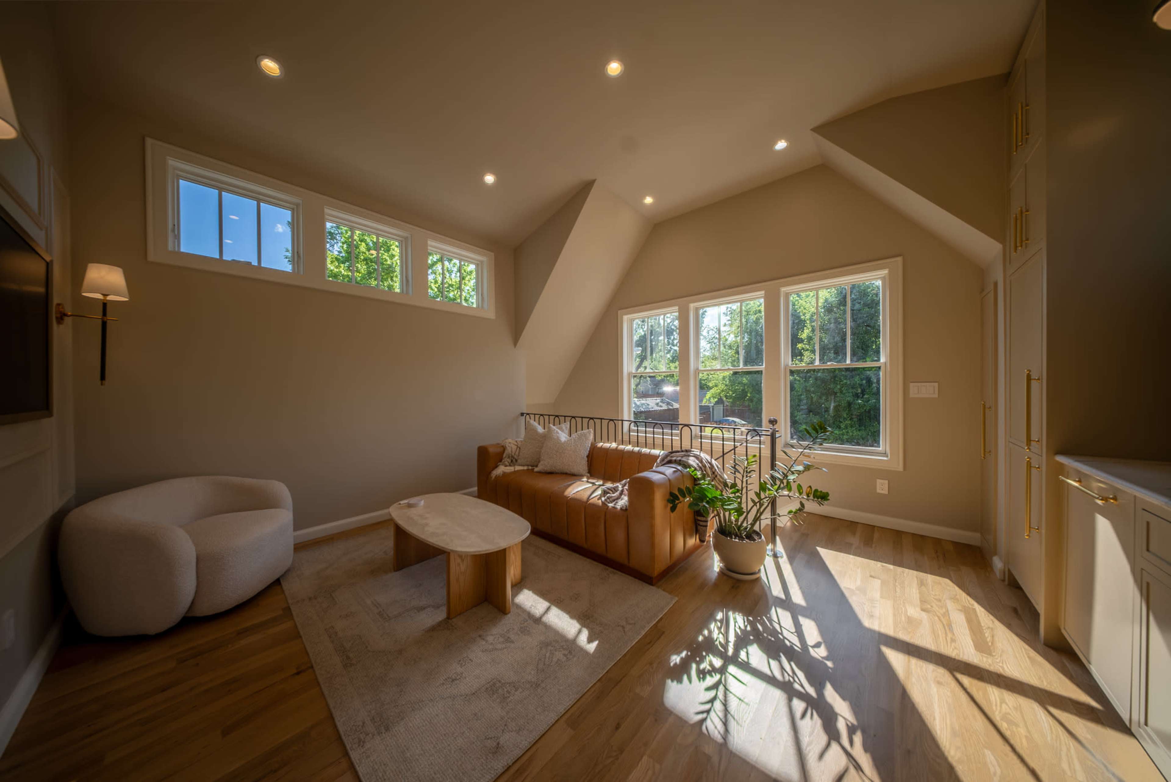 The image shows a modern living room with large windows, a brown leather sofa, a round coffee table, and natural wood flooring.