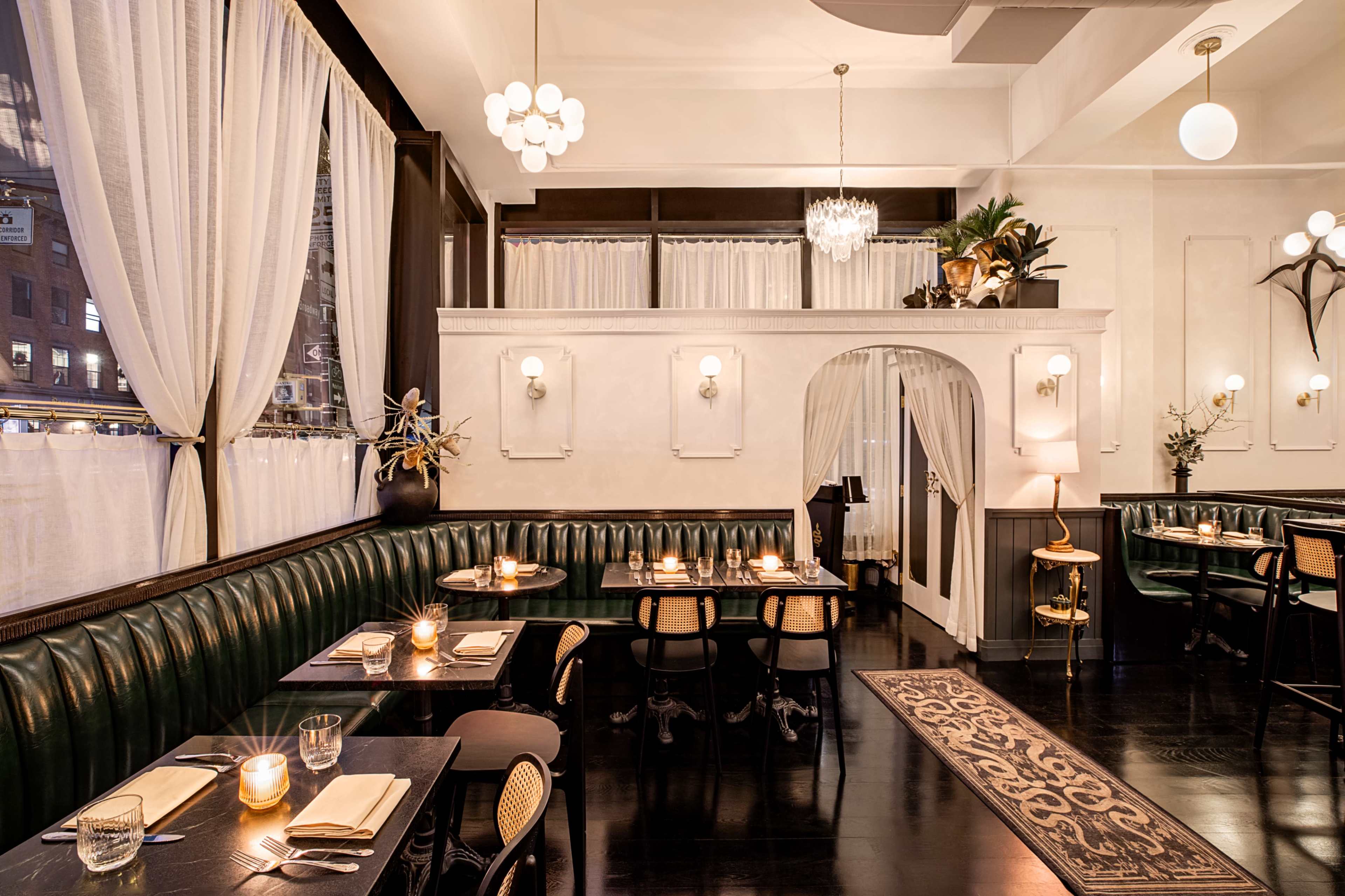 The image shows a stylish restaurant interior featuring green upholstered seating, round tables set with utensils, and large windows dressed with white curtains, illuminated by soft lighting.
