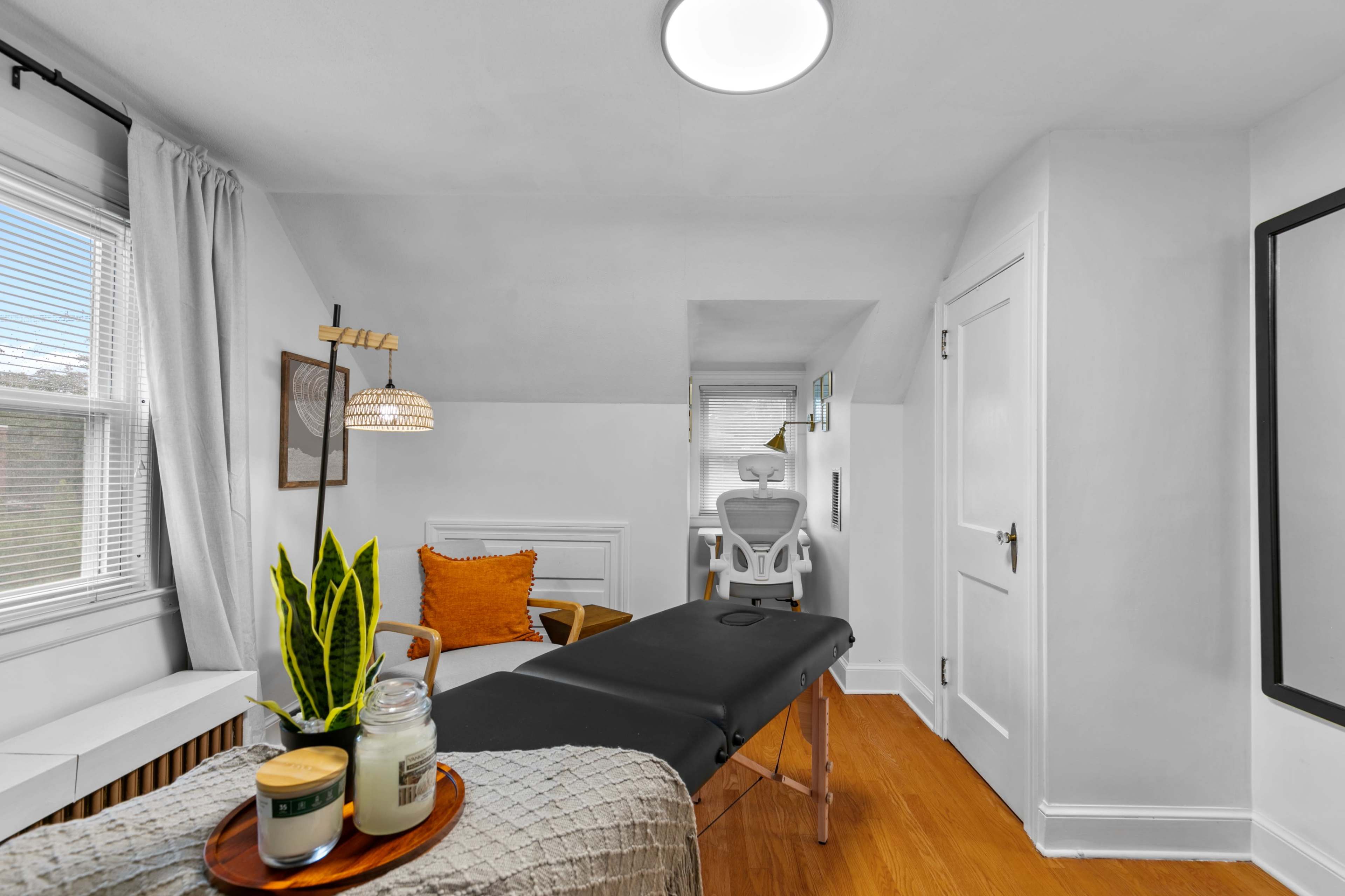 A well-lit therapy room featuring a treatment table, a chair, and decorative elements such as plants and candles.
