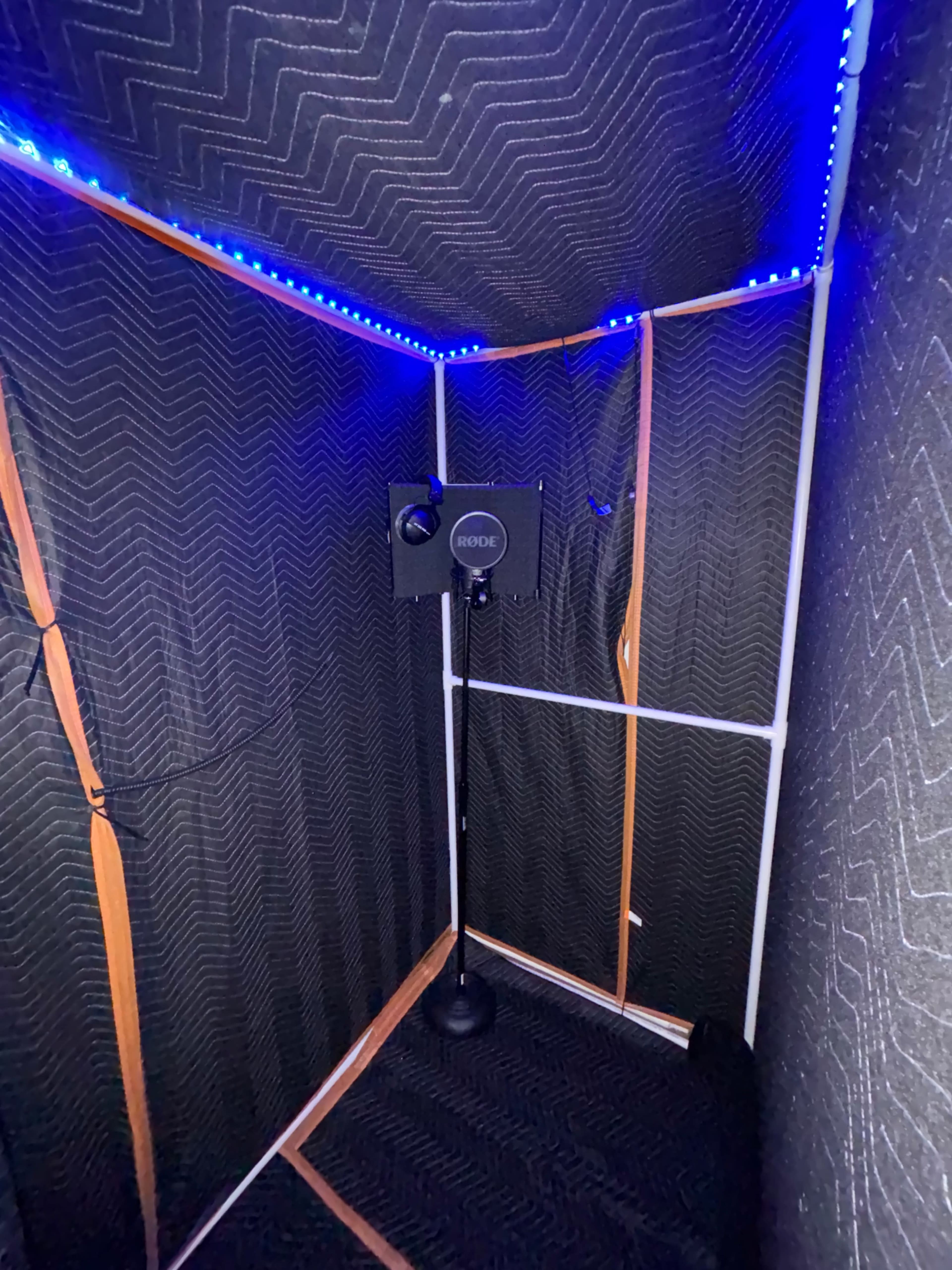 A soundproof booth lined with acoustic foam panels and illuminated by blue LED strip lights, featuring a microphone stand in the corner.