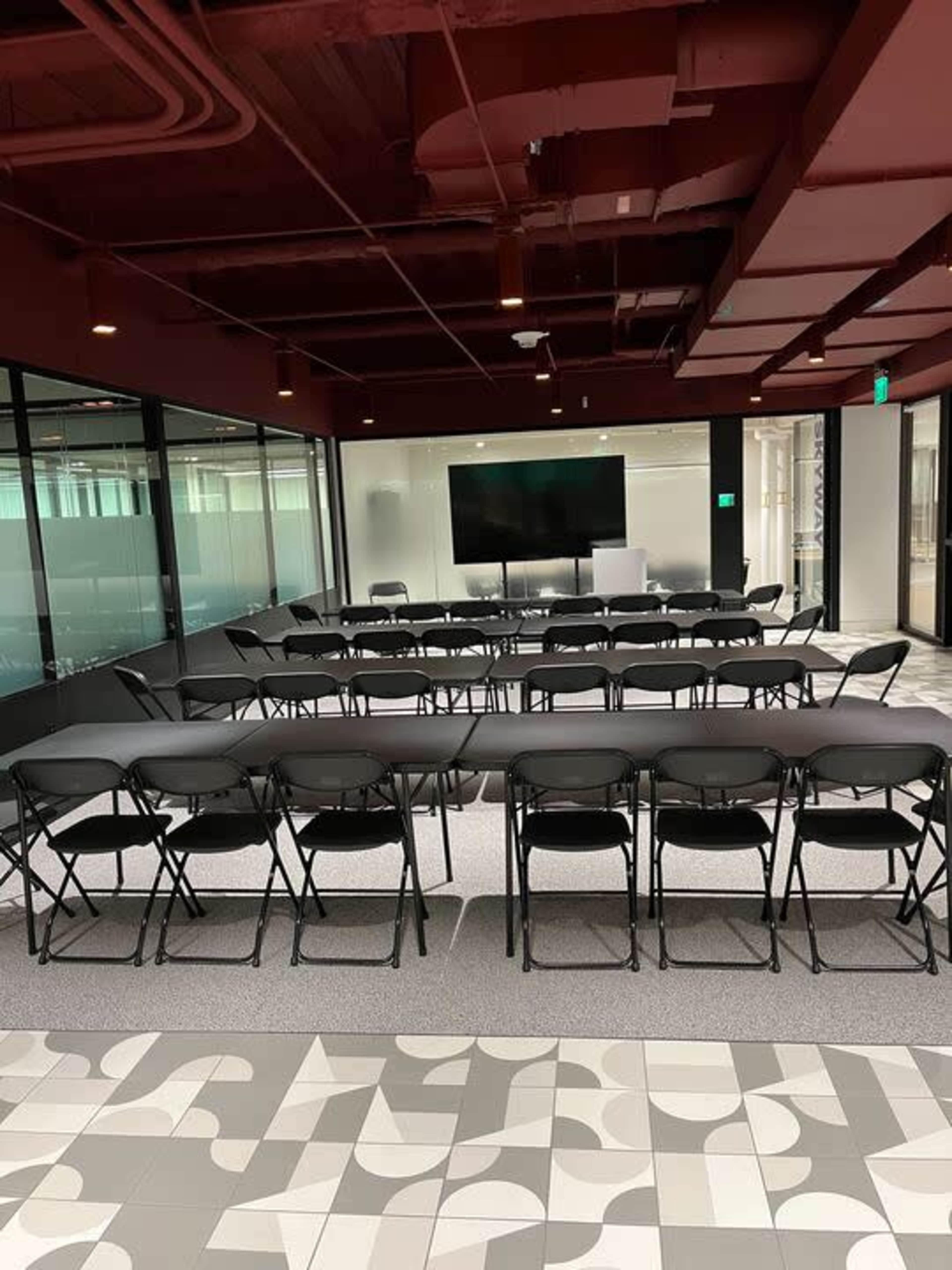 A large meeting room features a long black table surrounded by black folding chairs, with a large screen mounted on the wall at one end.