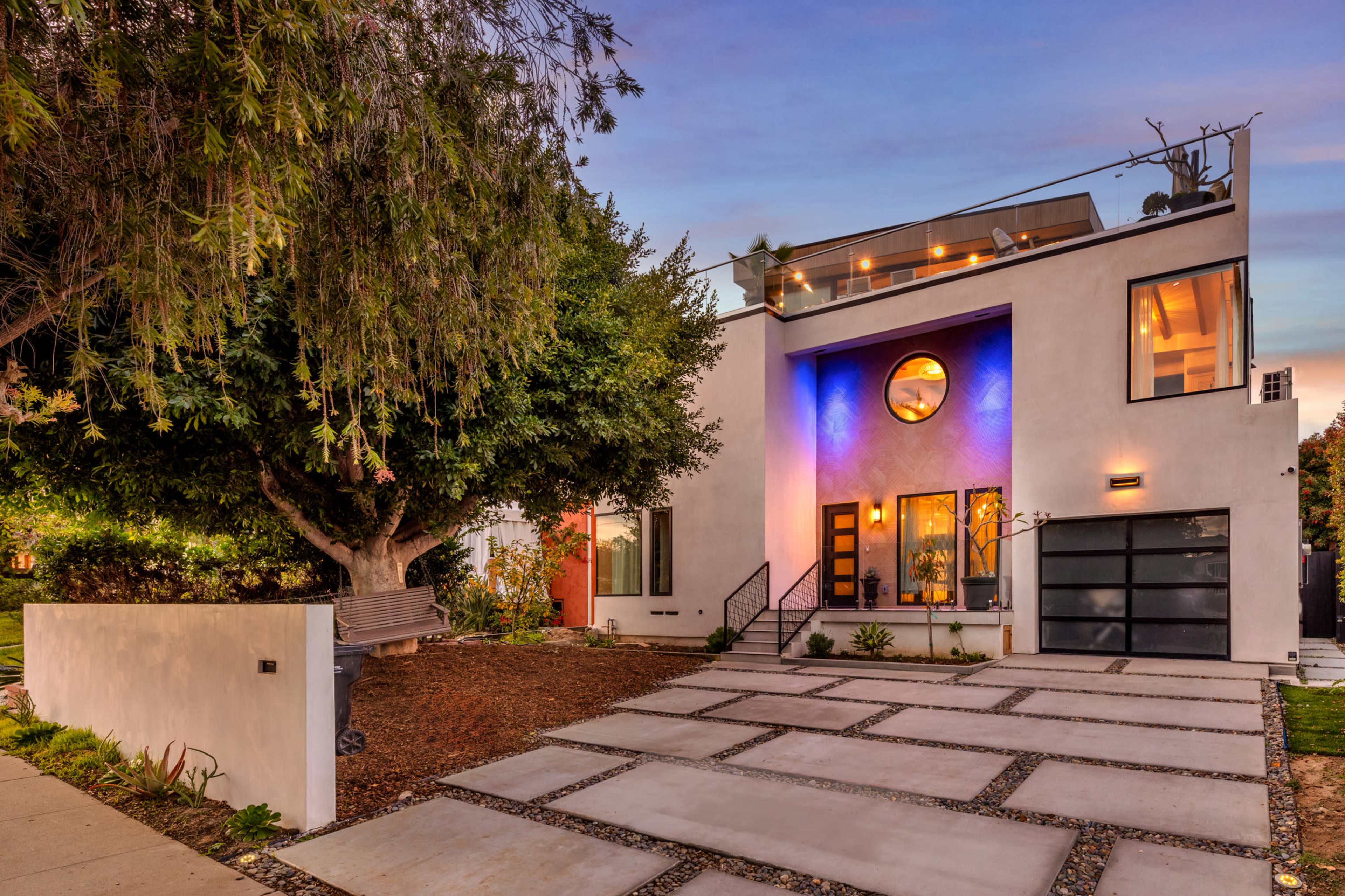 A modern two-story house features a curved window, a rooftop terrace, and a landscaped front yard with a stone walkway.