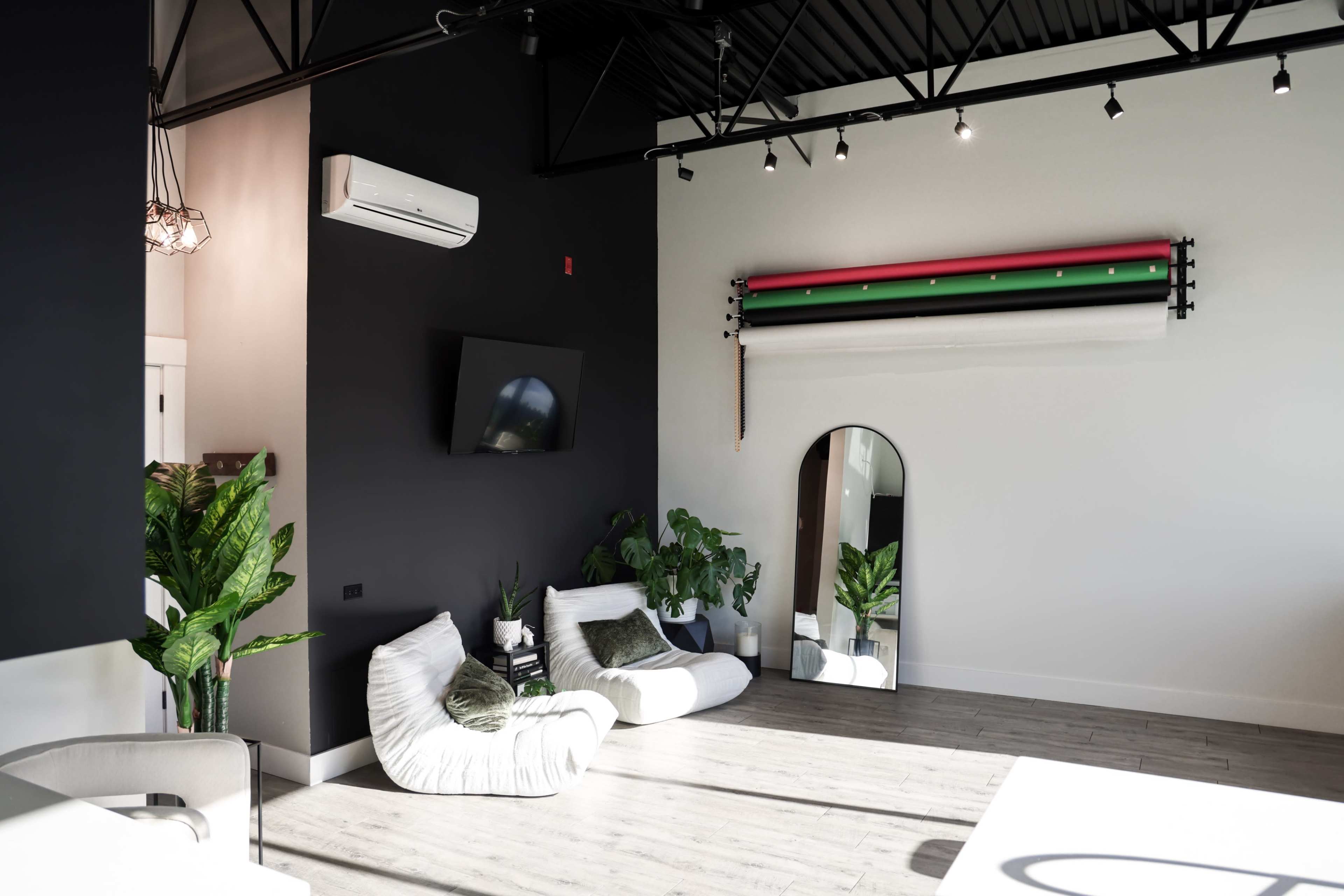 The image shows a modern indoor space with two white beanbag chairs, a large mirror, a wall-mounted television, and plants against a backdrop of black and white walls with colorful horizontal bars.
